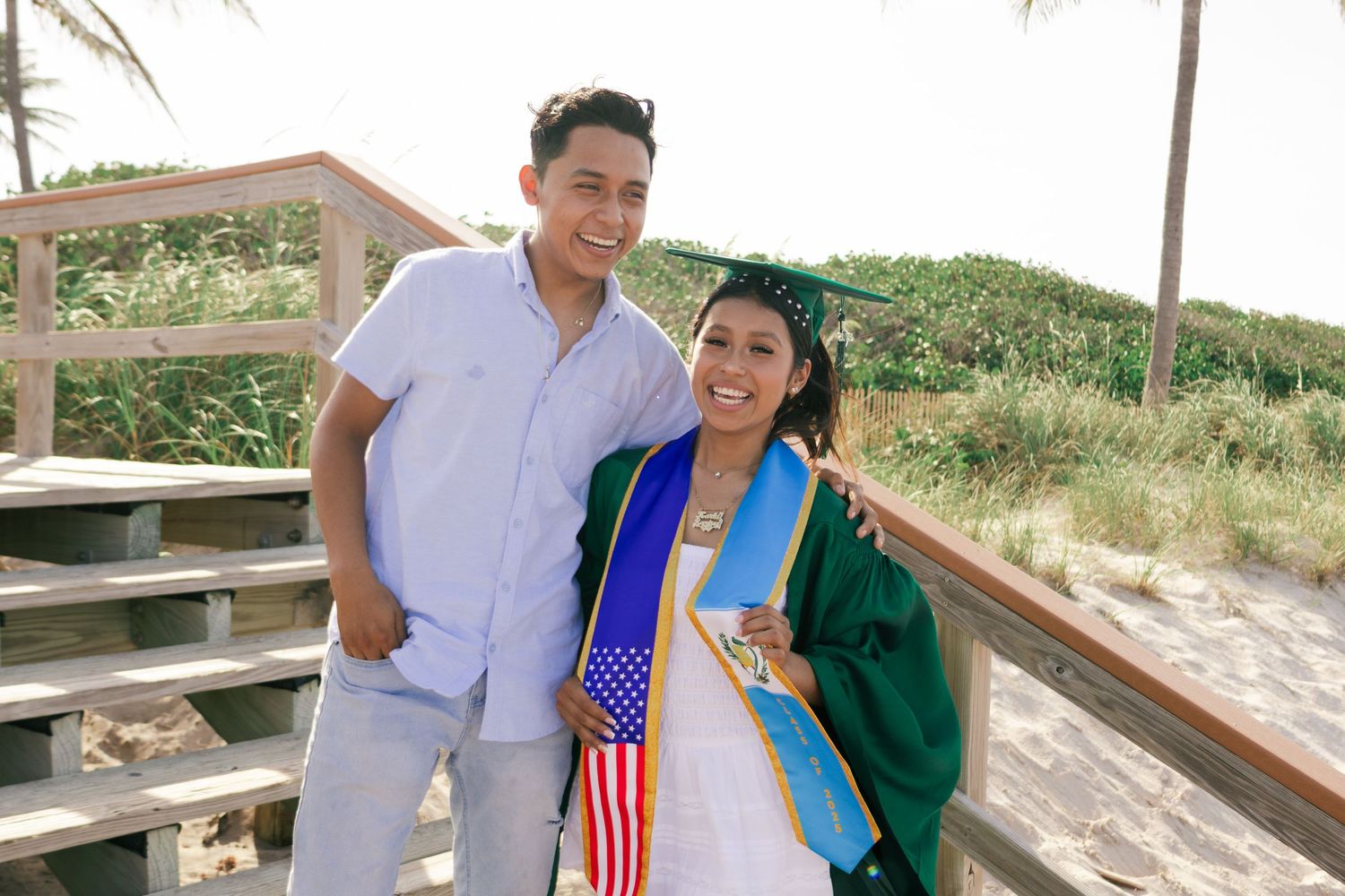 Graduate in green cap and gown poses with a companion on a sunny beach boardwalk holding an American flag.