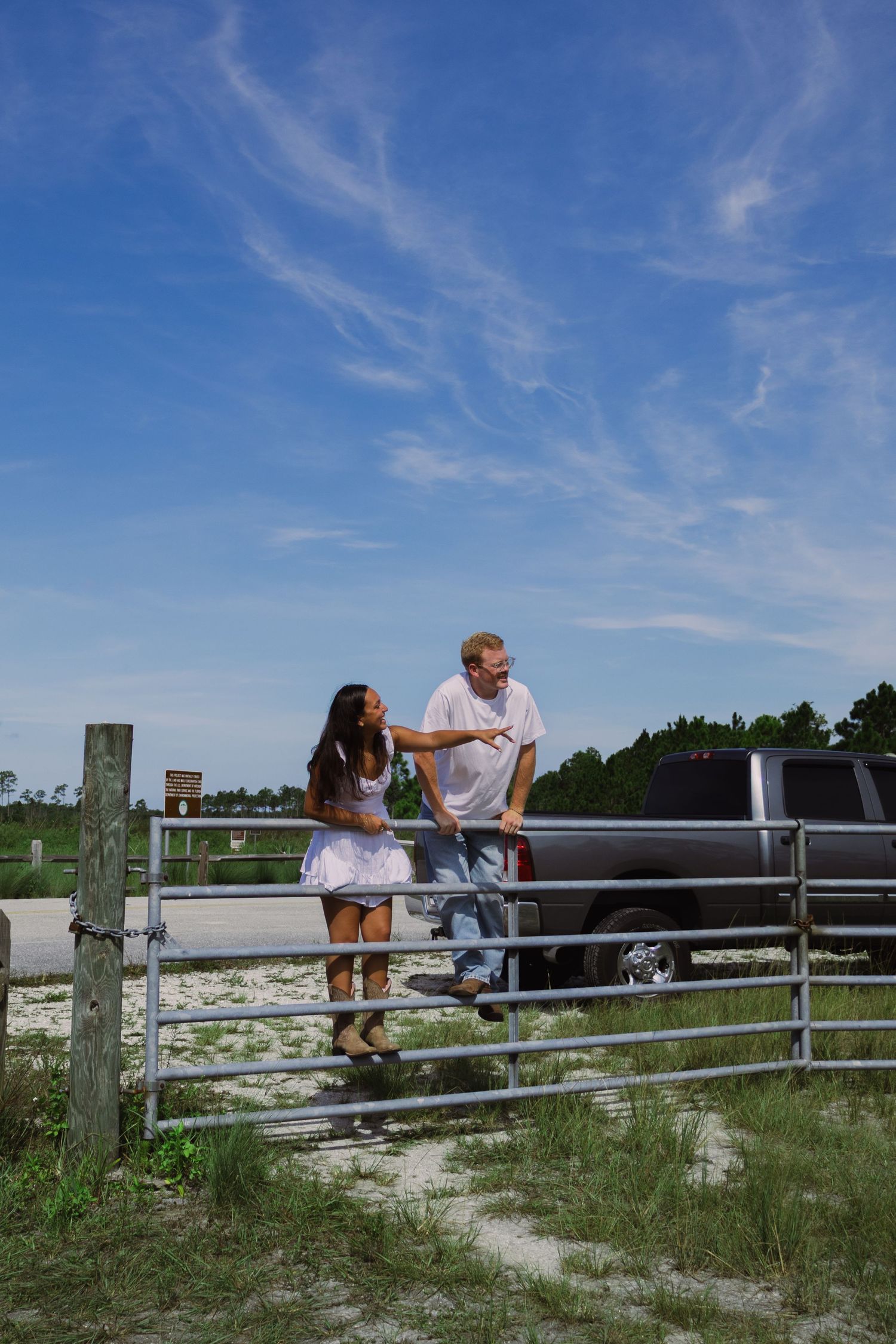 Couple talking and leaning on a metal farm gate with a pickup truck and blue sky in the background.