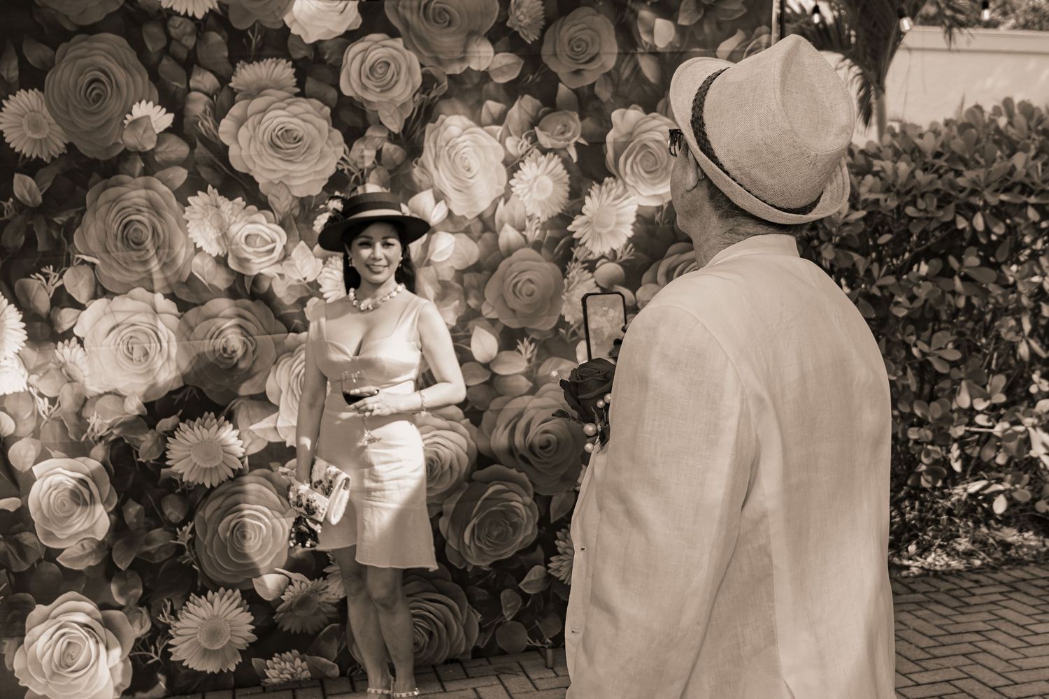 Black and white photo of woman posing at floral backdrop while man photographs her at Derby party.