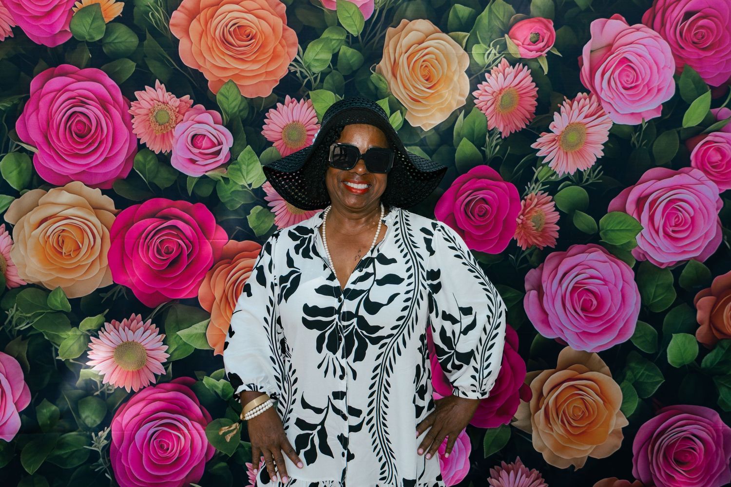 Woman in black and white floral dress posing in front of a colorful rose flower wall backdrop.