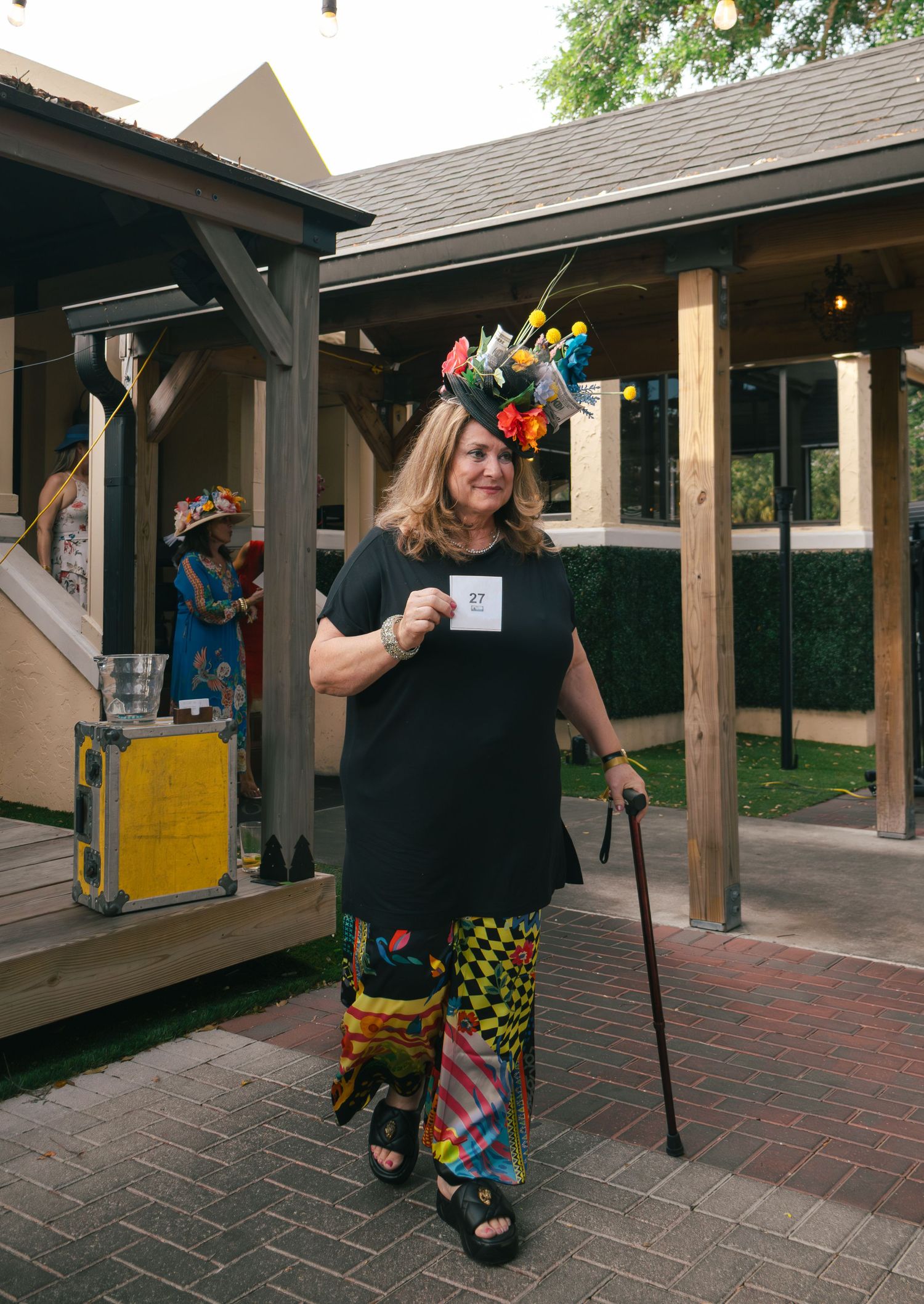 Woman with colorful floral headpiece and patterned pants holds card at outdoor Derby event.