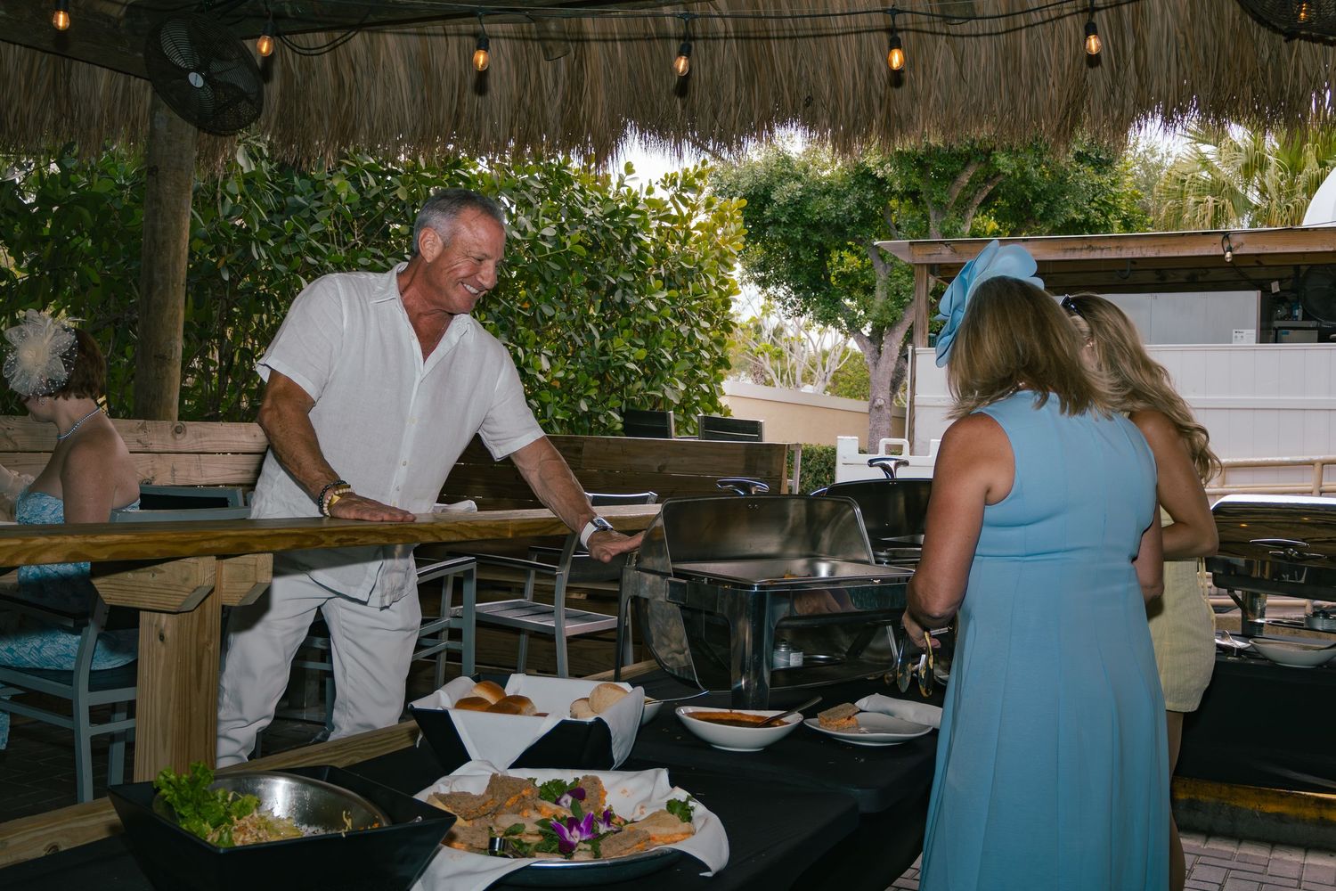 Chef in white serving food at outdoor buffet station to guest in blue hat.