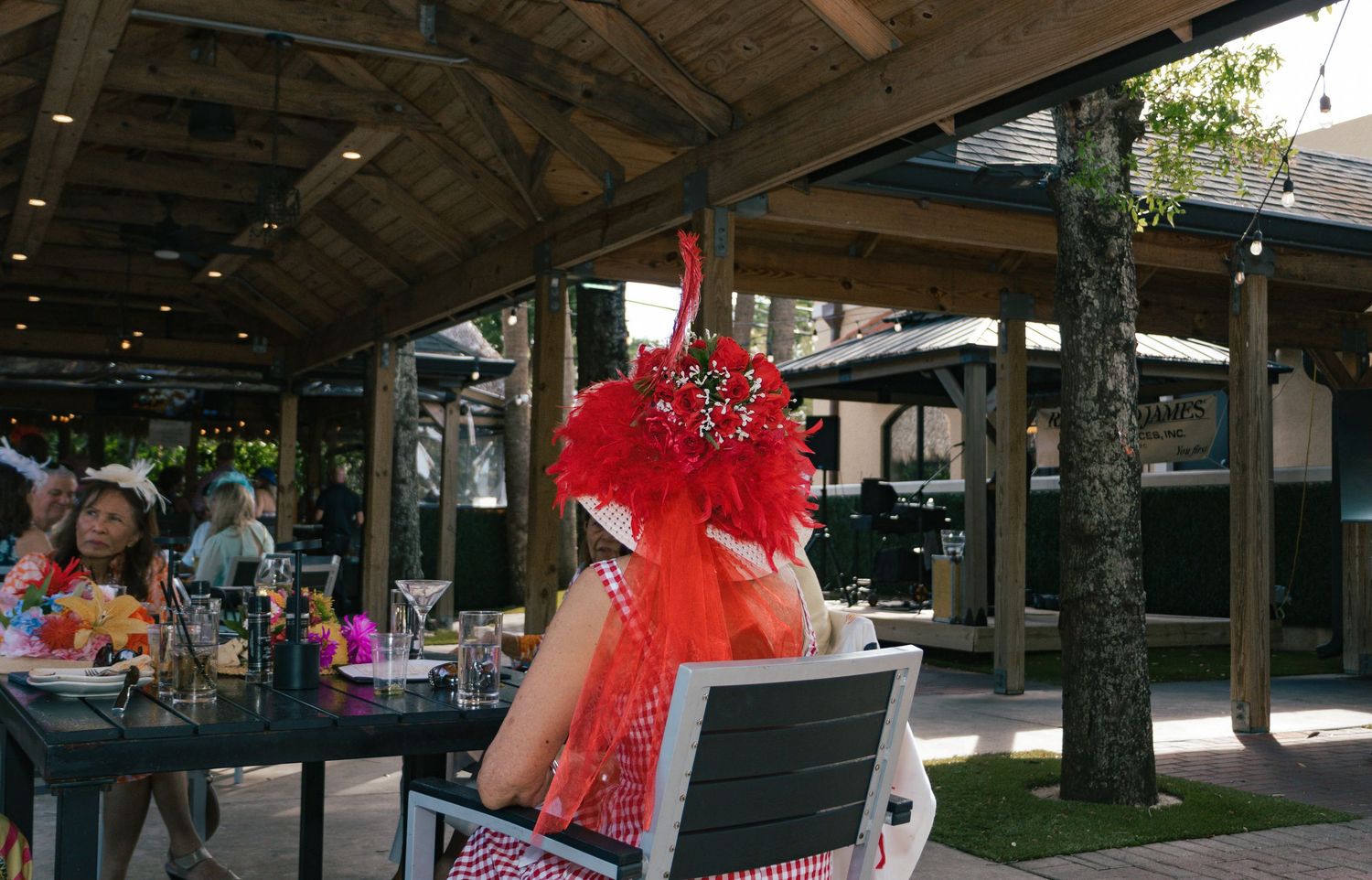 Woman in red gingham dress and large red feathered hat seated at an outdoor Kentucky Derby party.