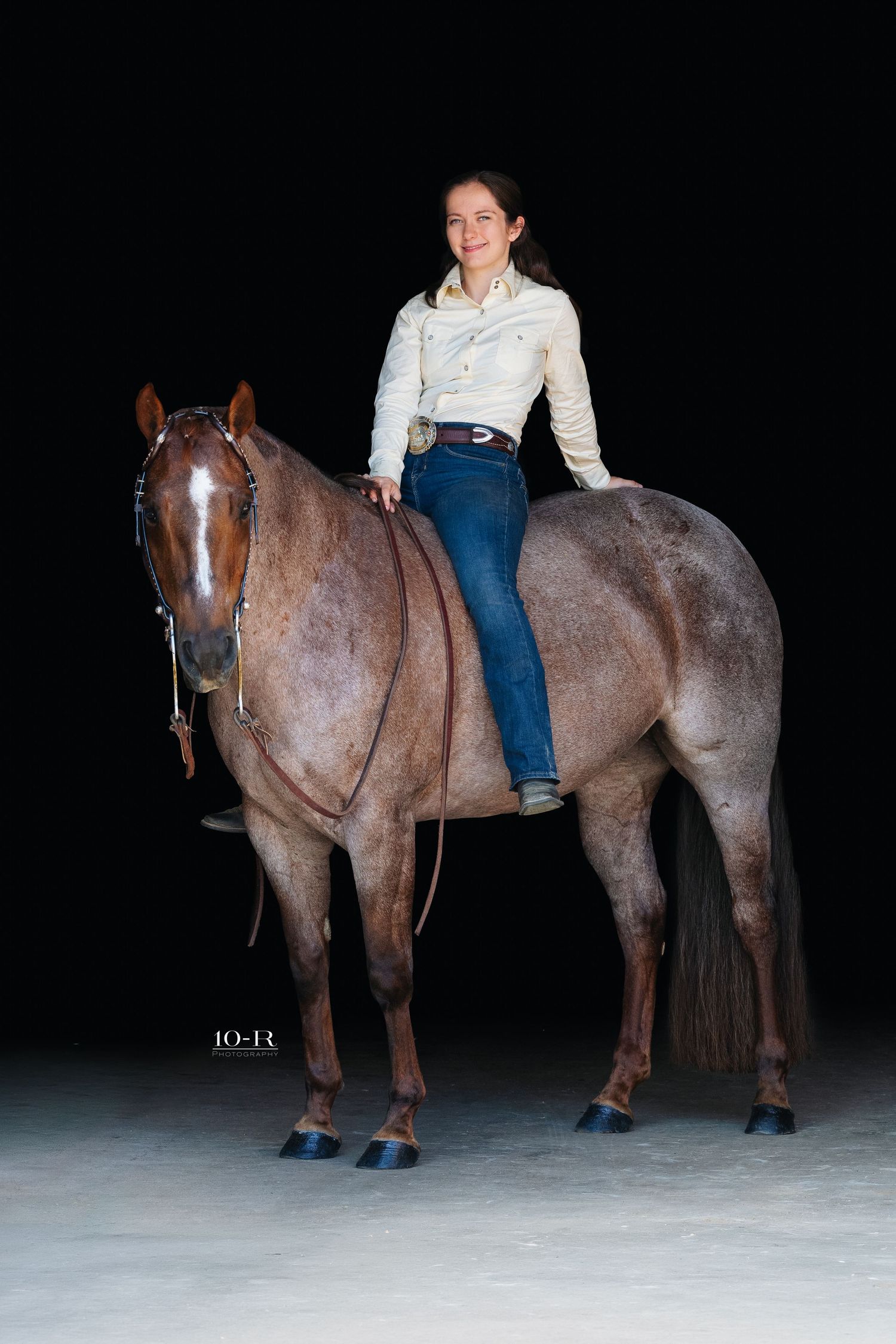 Young woman in white shirt and jeans riding a roan horse against a black background.