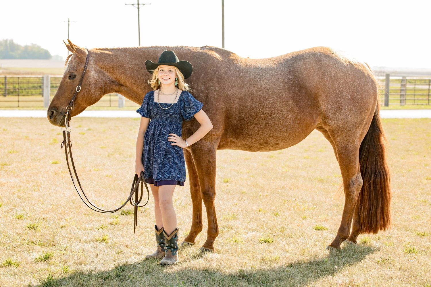 A brown horse stands beside someone in a blue dress and cowboy boots in a sunny field.