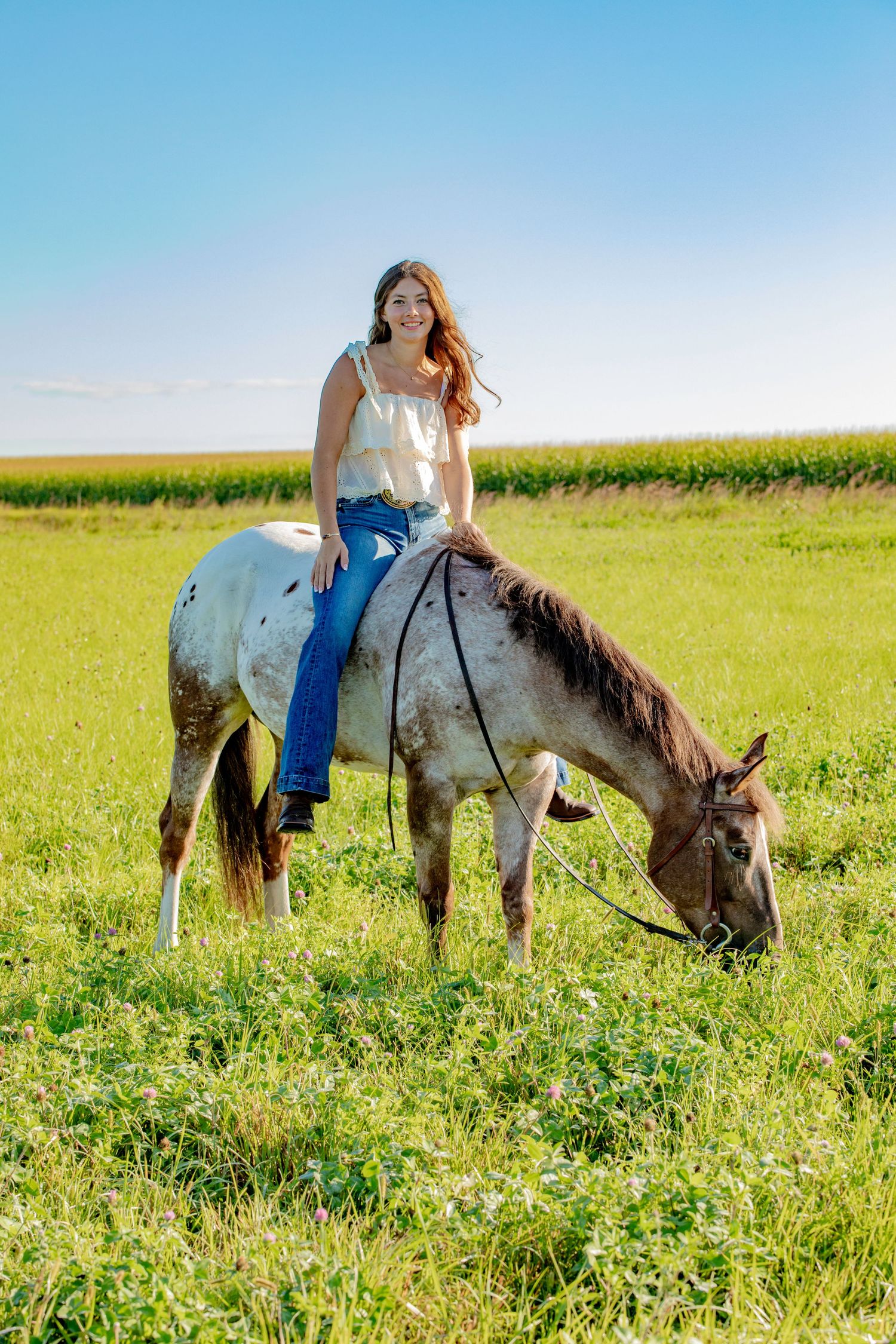 A painted horse grazes in a green field while being ridden on a sunny summer day.