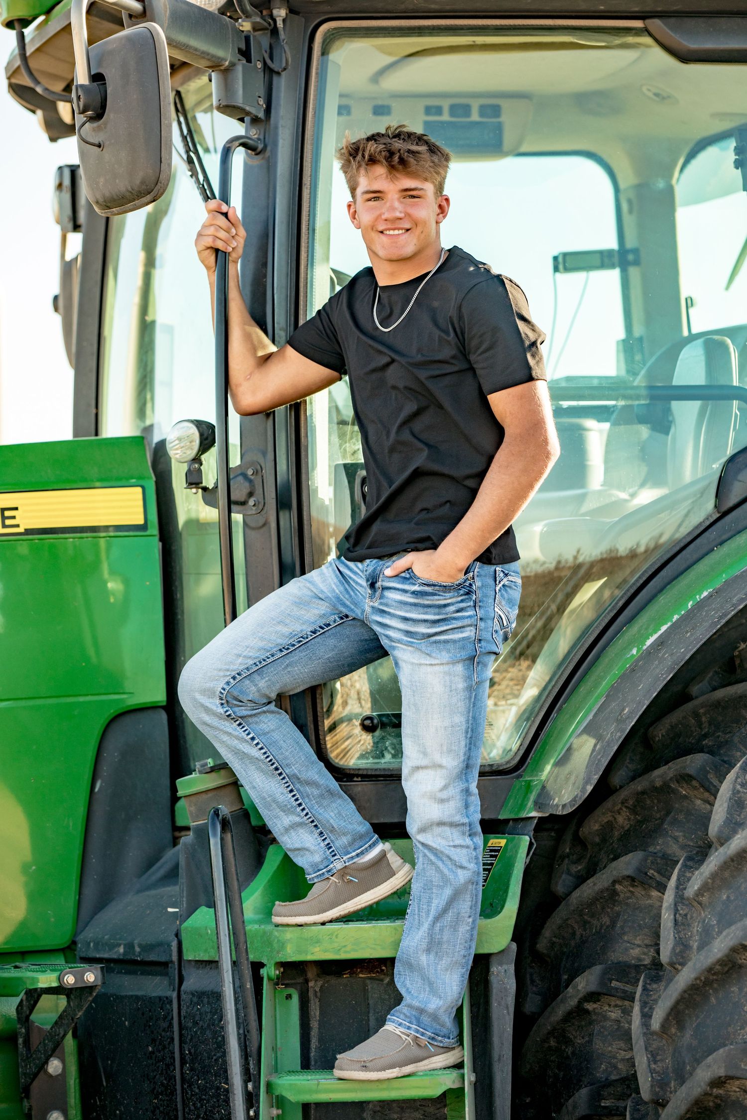 Person in black t-shirt and jeans poses on steps of green John Deere tractor.
