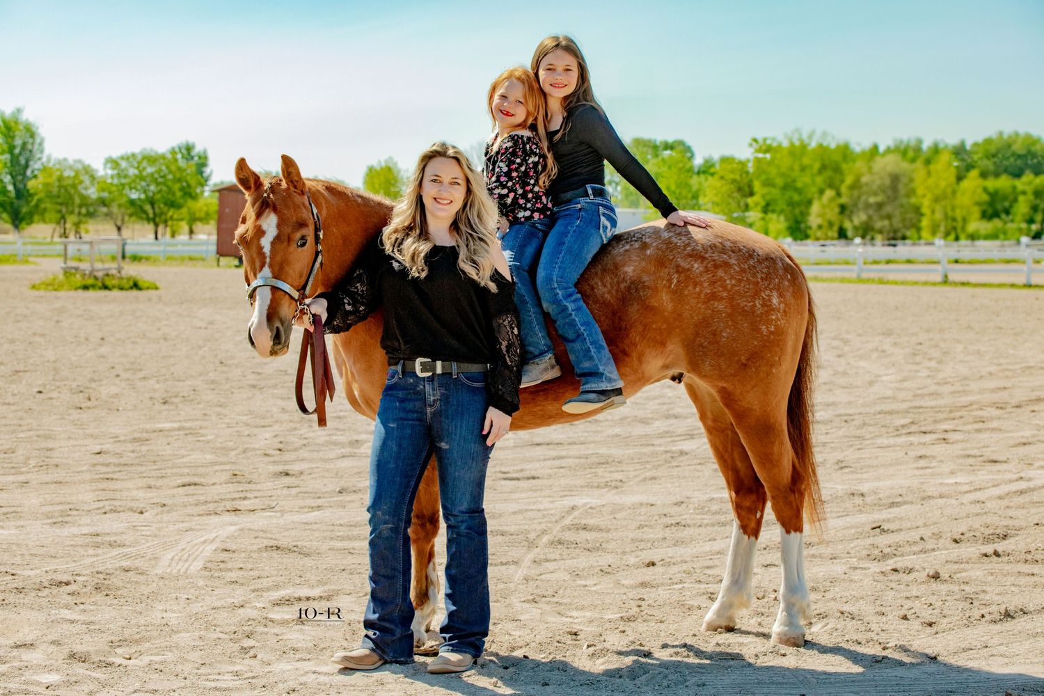 A brown horse stands with riders at an outdoor riding arena with green trees visible in the background.