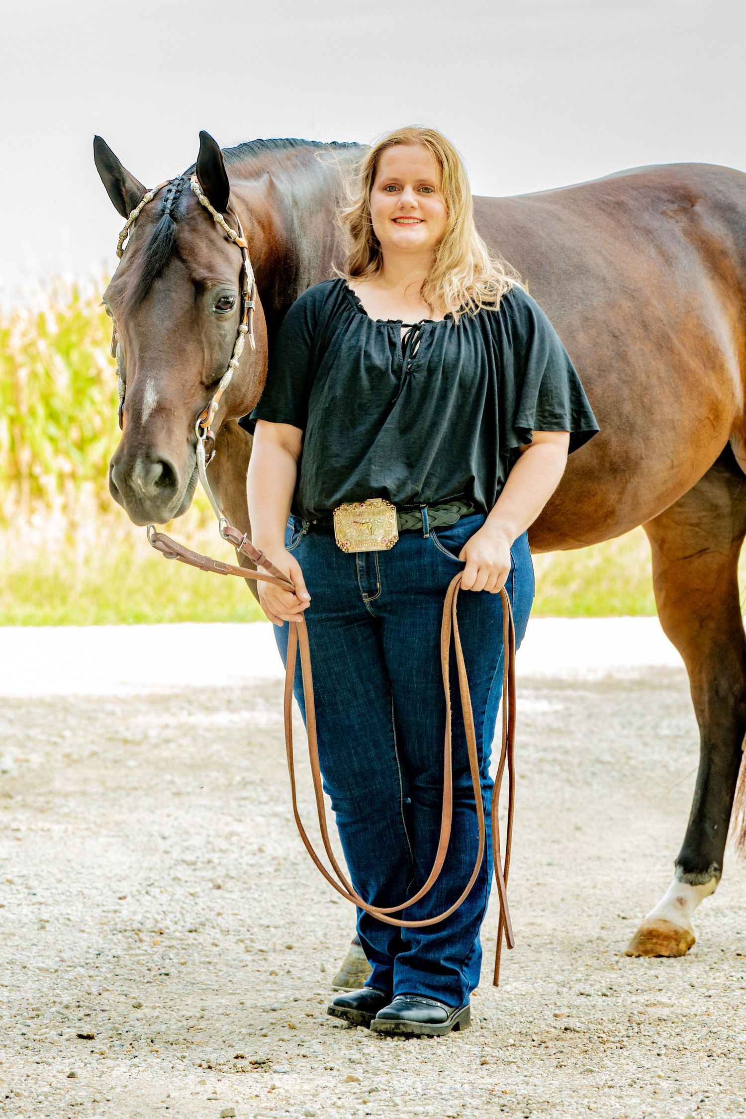 Blonde woman in black top and jeans holds leather reins beside a bay horse outdoors.