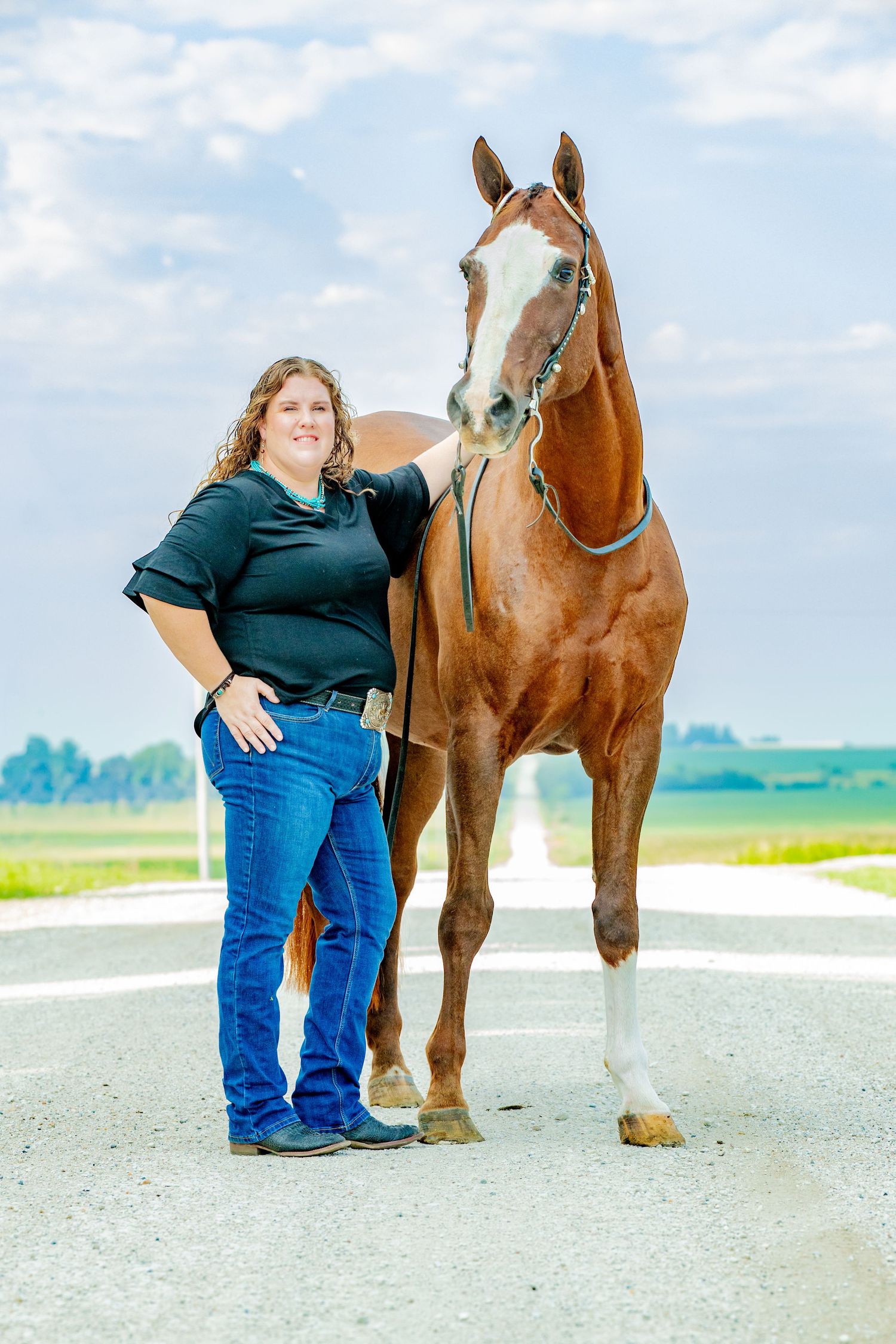 A smiling woman in a black shirt and jeans stands beside a chestnut horse on a rural road under a cloudy sky.