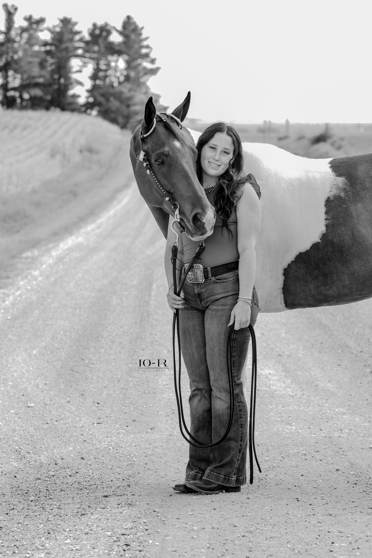Black and white photo of a young woman hugging a paint horse on a rural dirt road with trees in the background.