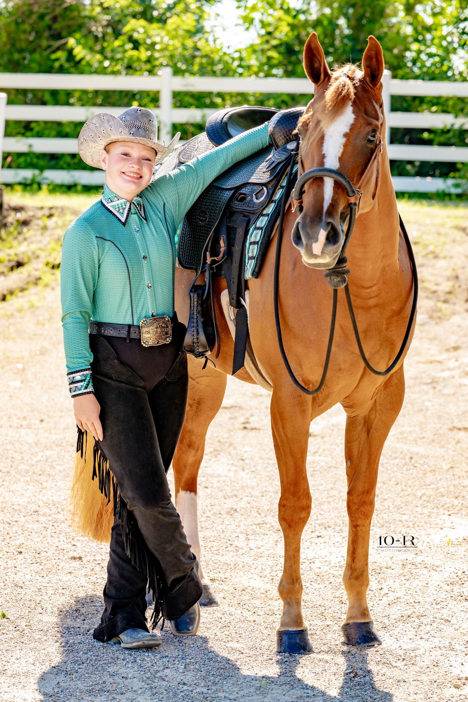 Young cowboy in green shirt and chaps poses proudly beside a saddled chestnut horse at an equestrian facility.