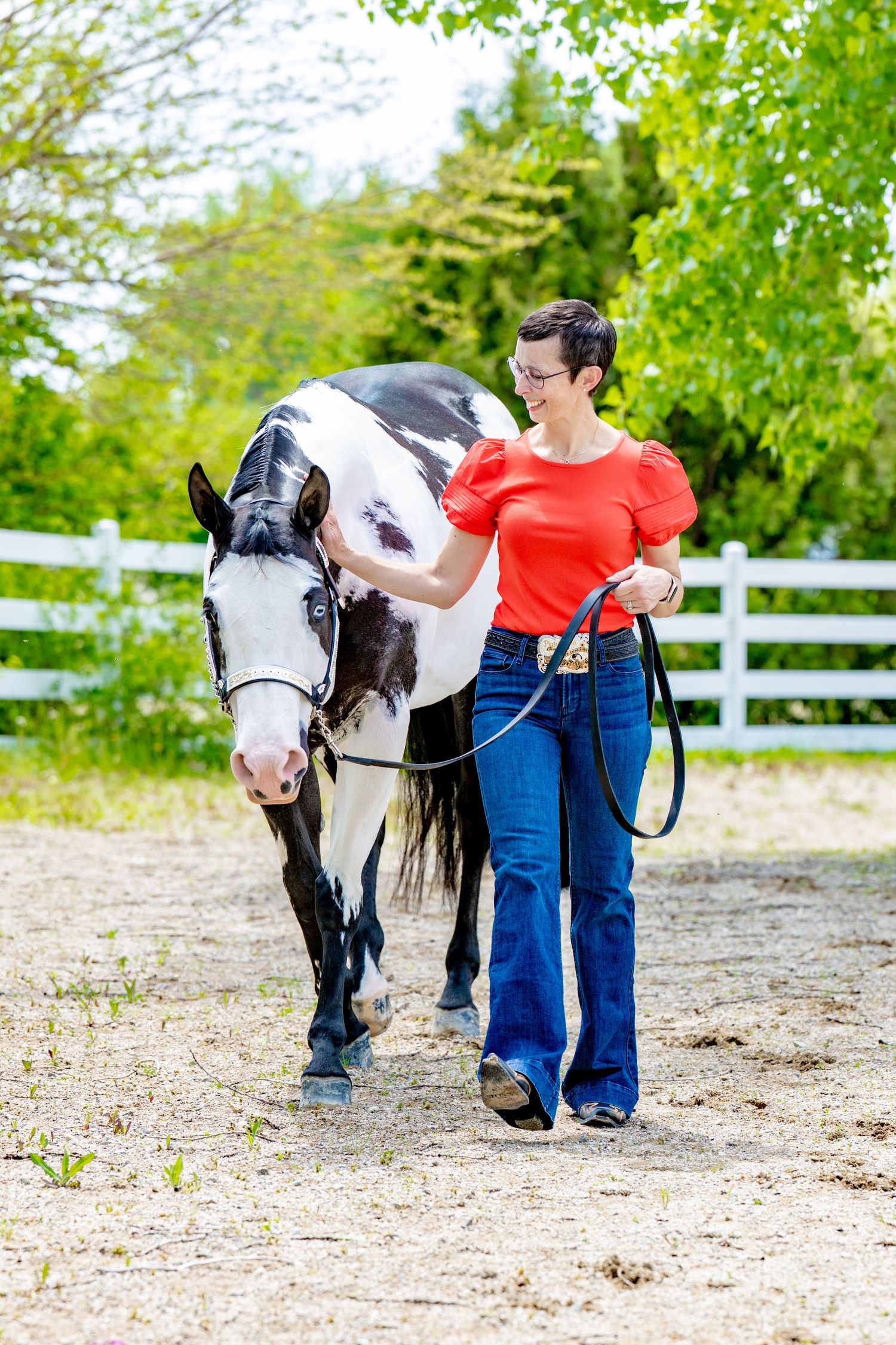 Woman in red top and jeans leading a black and white paint horse in a sandy paddock with white fence.