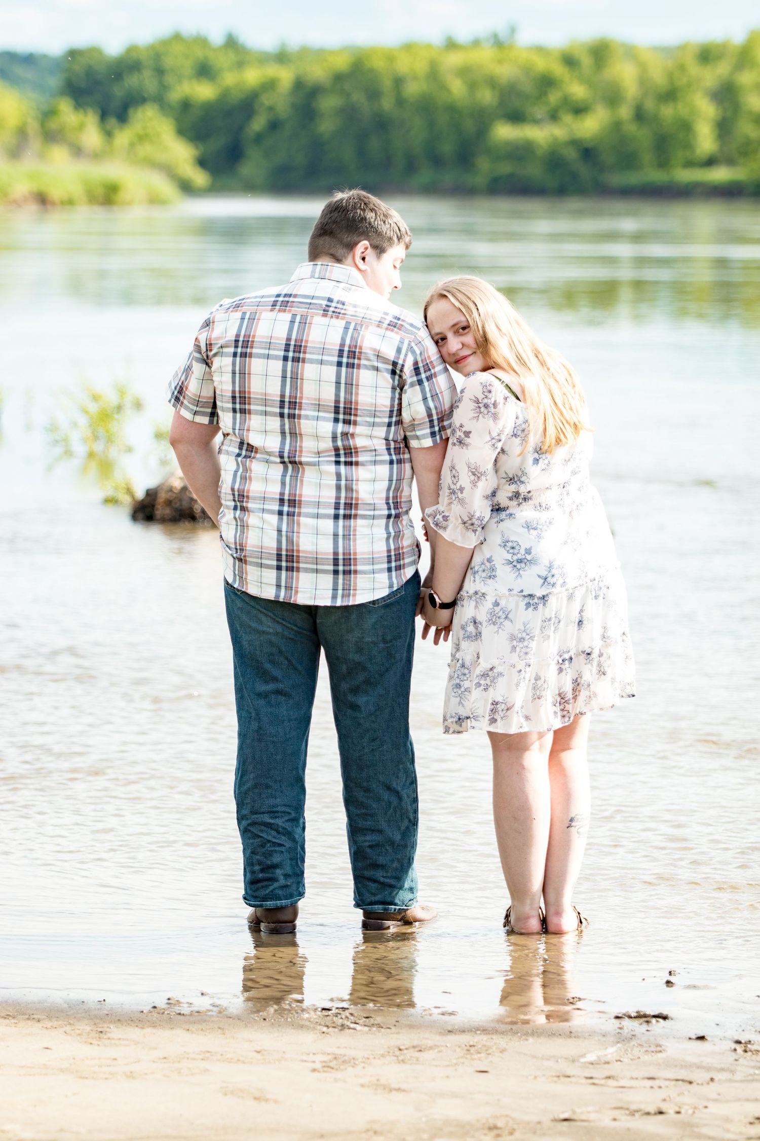 A couple stands barefoot in shallow water by a lake on a sunny day, wearing casual summer attire.