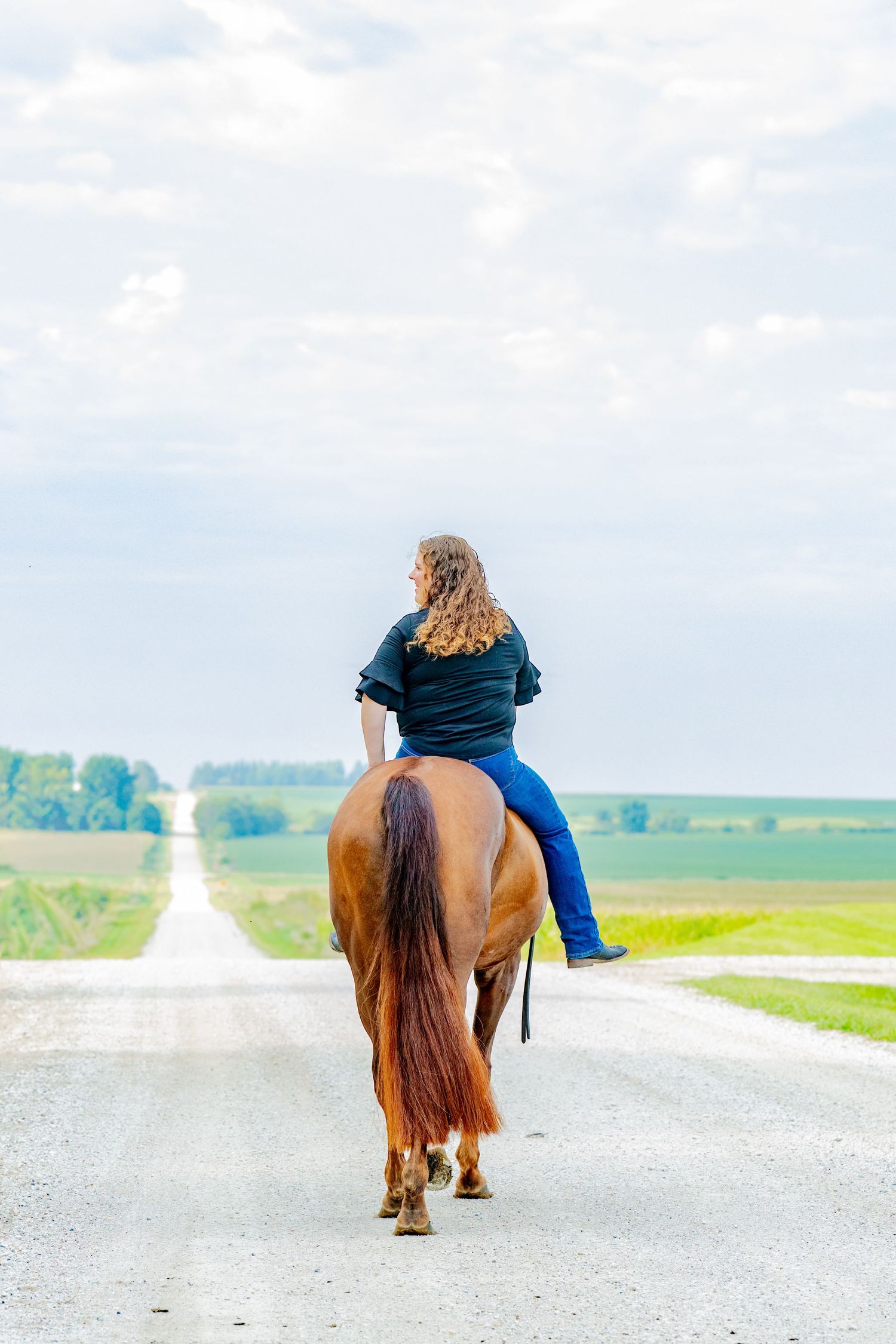Young woman with curly hair riding a chestnut horse down a rural gravel road surrounded by green fields.