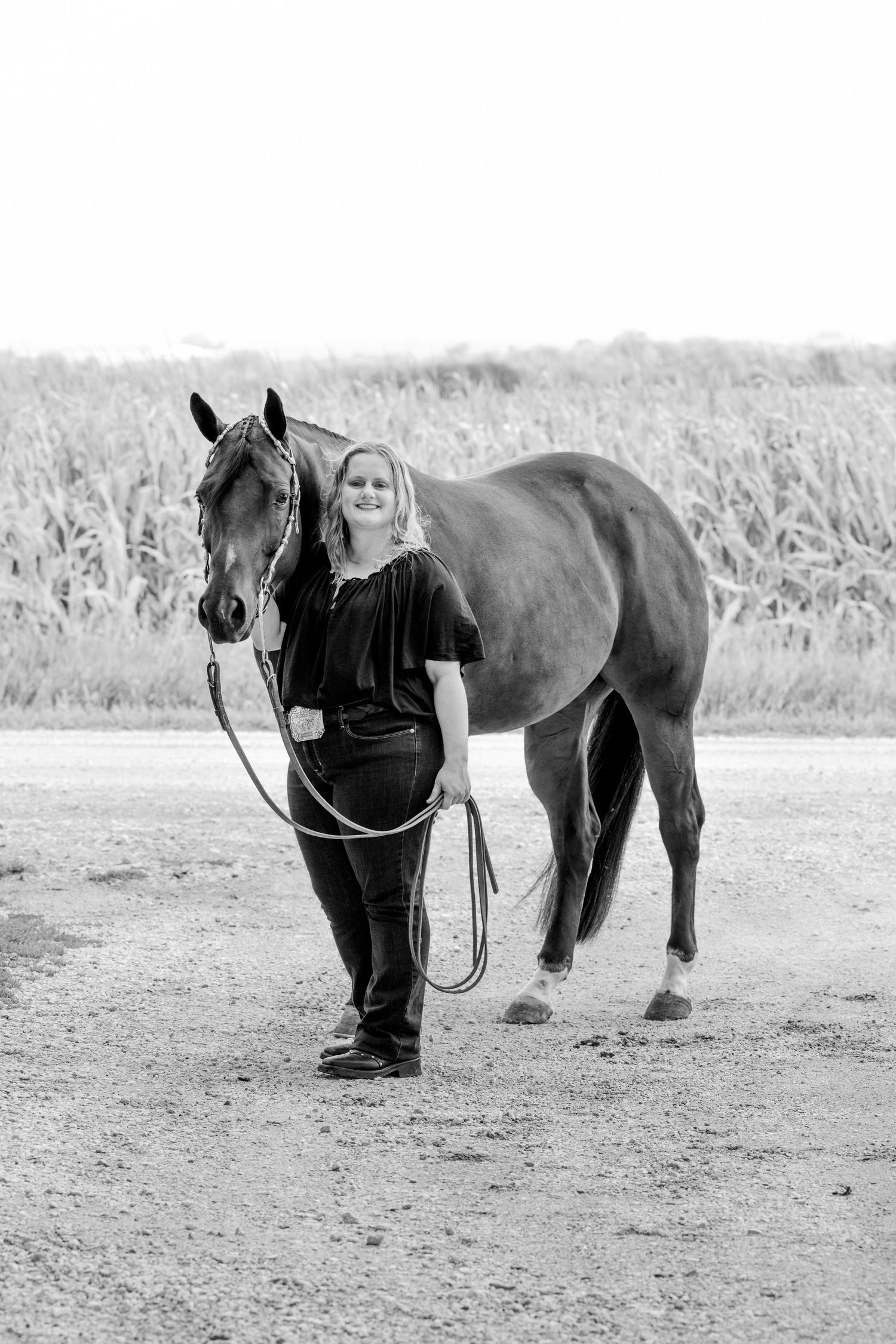 Black and white photo of a smiling woman standing beside a horse near a cornfield, holding a lead rope.
