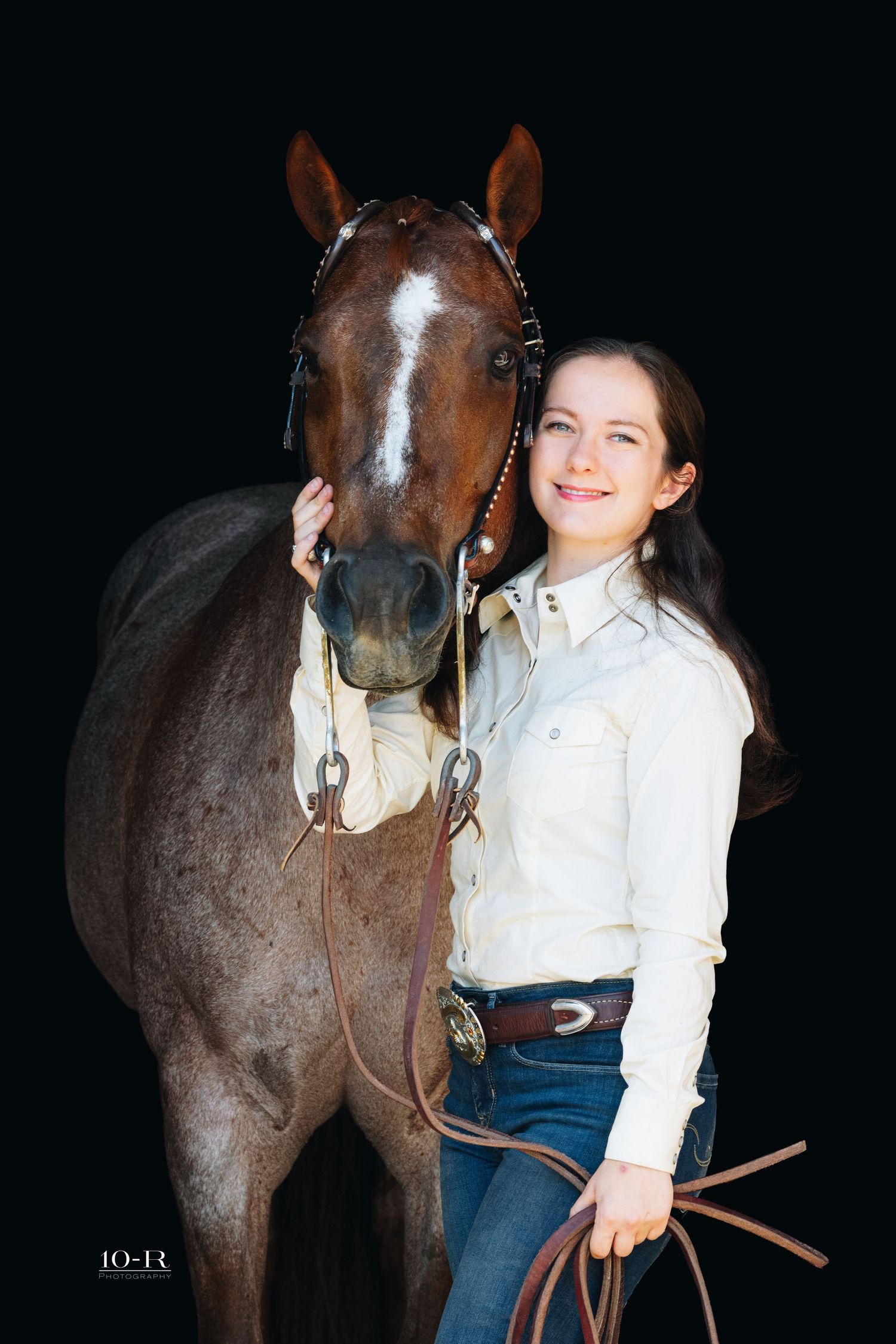 Young woman in western attire smiling beside a dark bay horse against a black studio background.