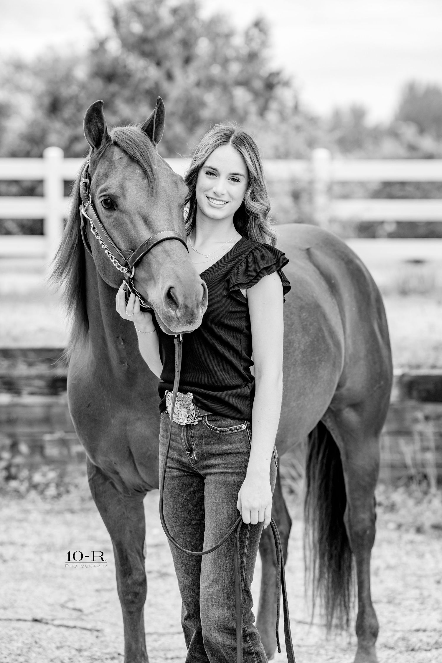 Young woman smiling beside a horse in a fenced arena, wearing jeans and a black top, black and white photo.