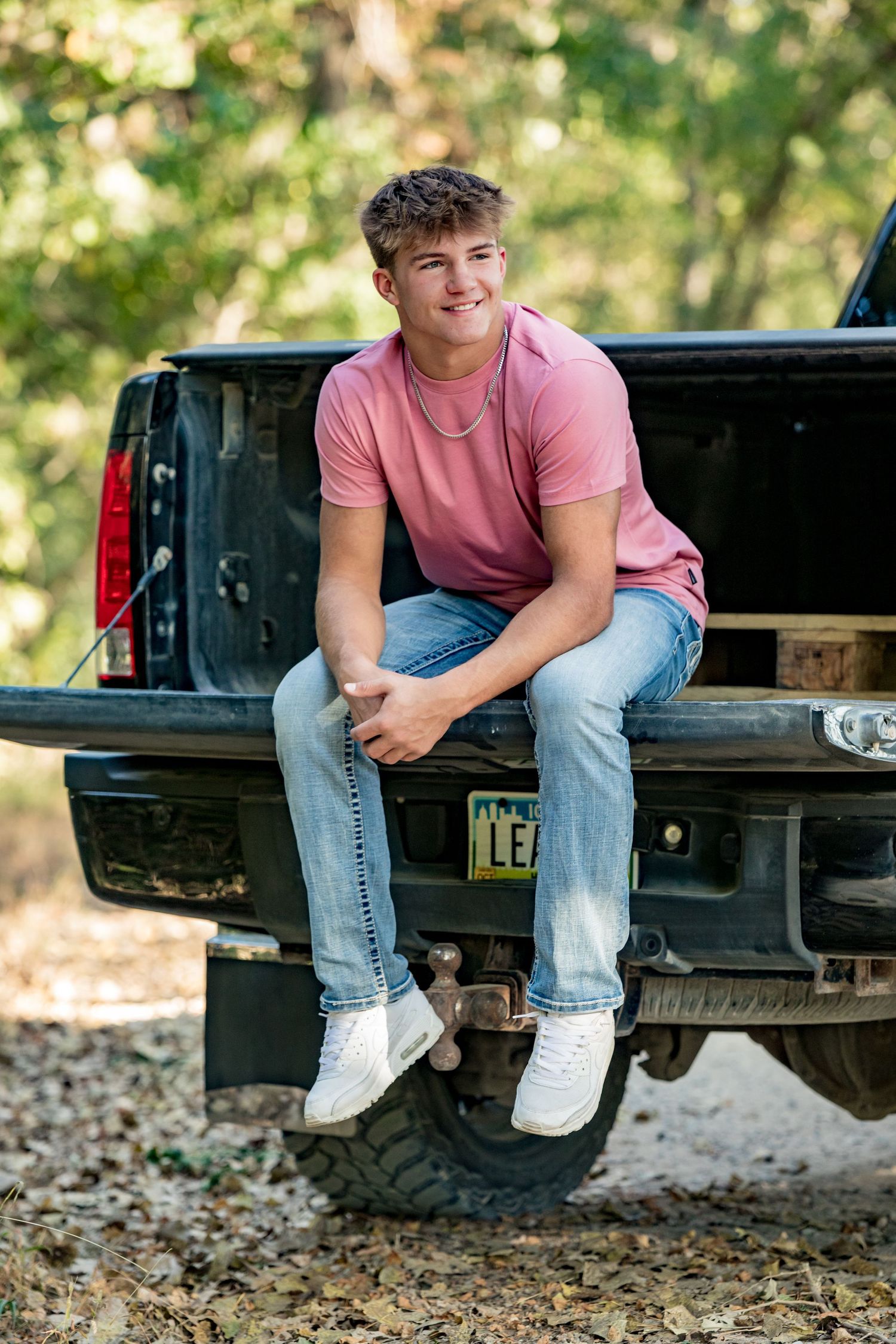 A person in a pink t-shirt and jeans sits casually on the tailgate of a black pickup truck in an outdoor setting.