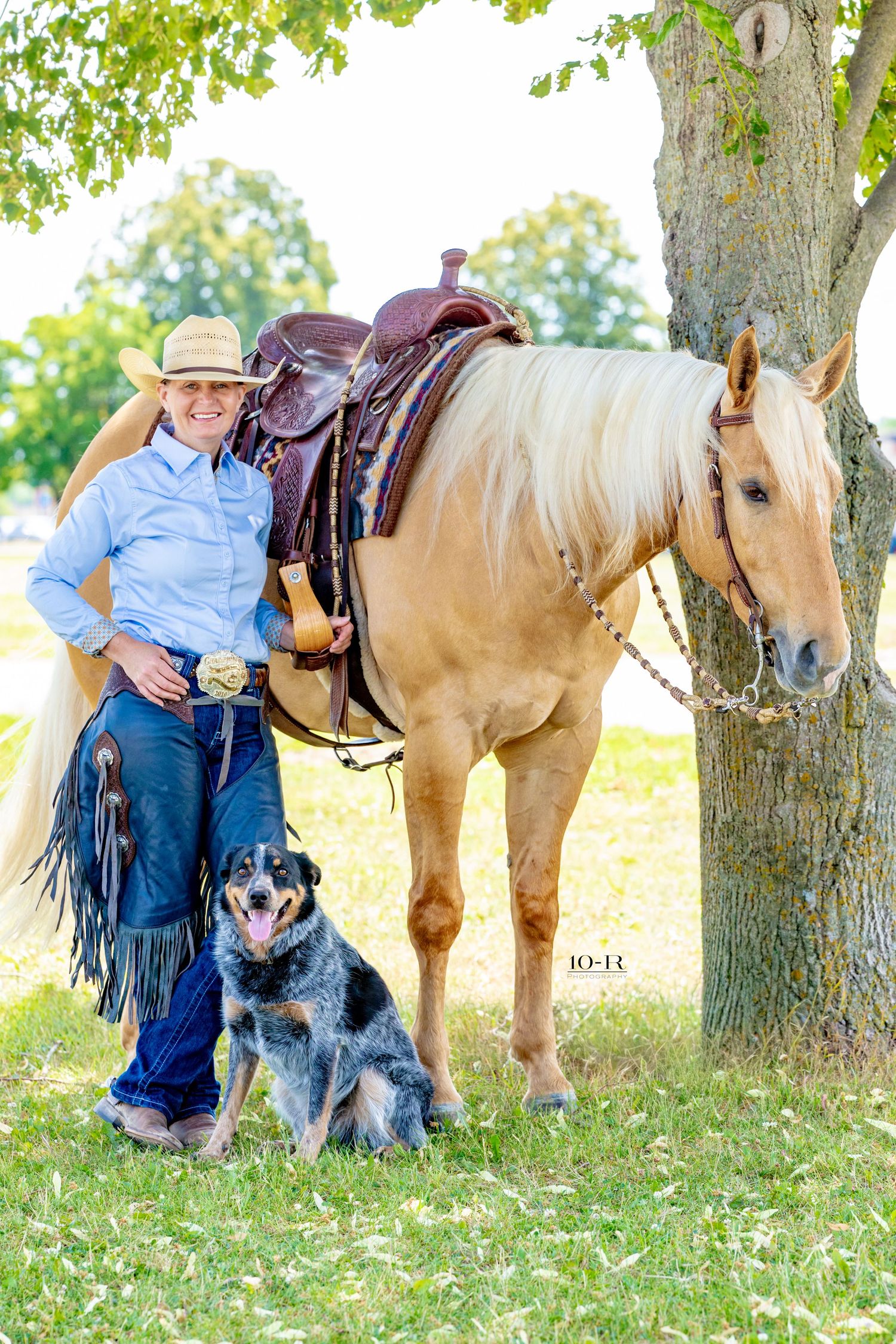 Cowgirl in hat and fringe chaps poses with palomino horse and blue heeler dog under a tree outdoors.