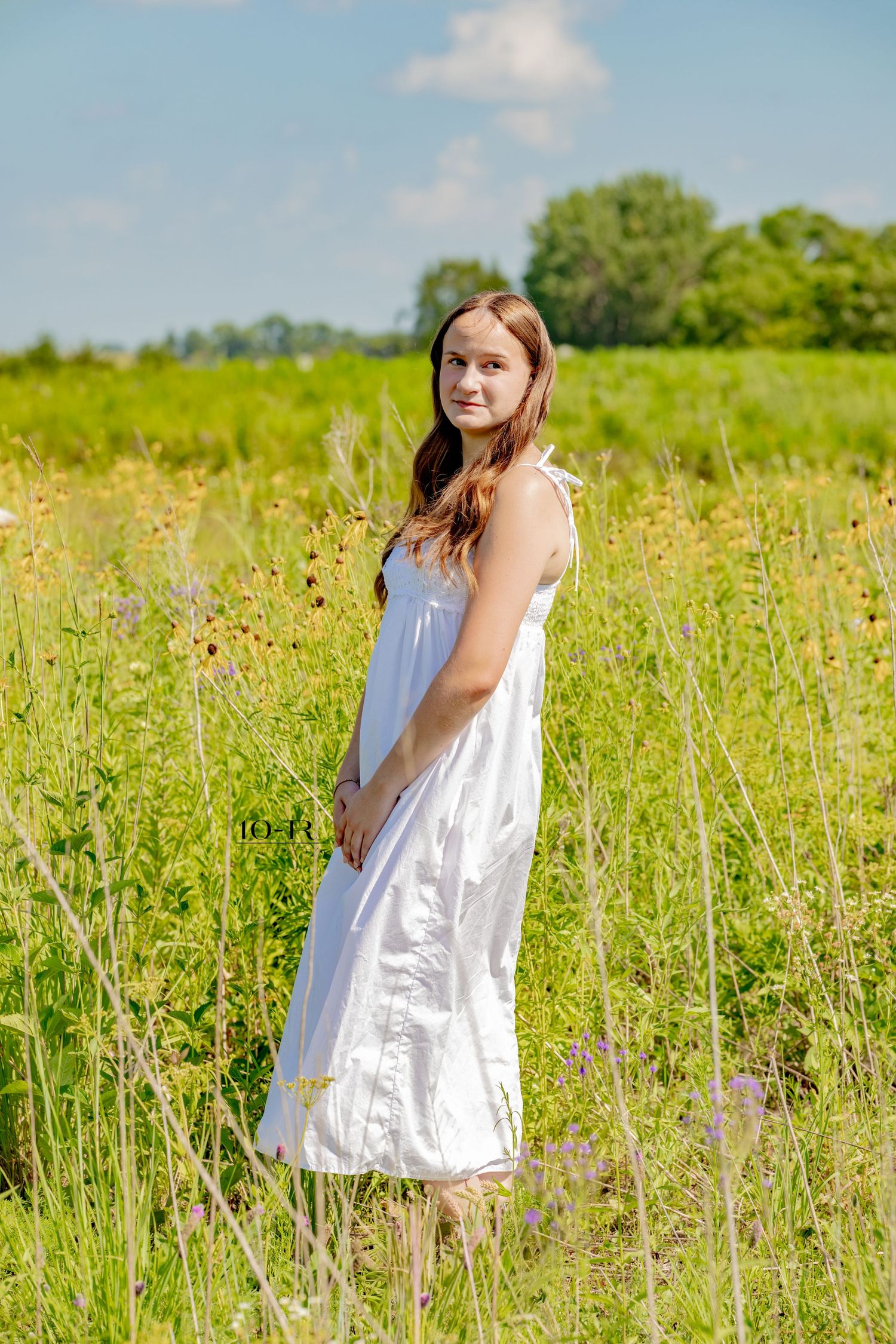 Young woman in white sundress standing in a wildflower meadow on a sunny summer day.
