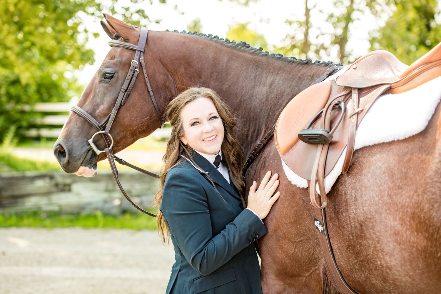 Rider in navy show jacket poses with bay horse wearing English tack outdoors.