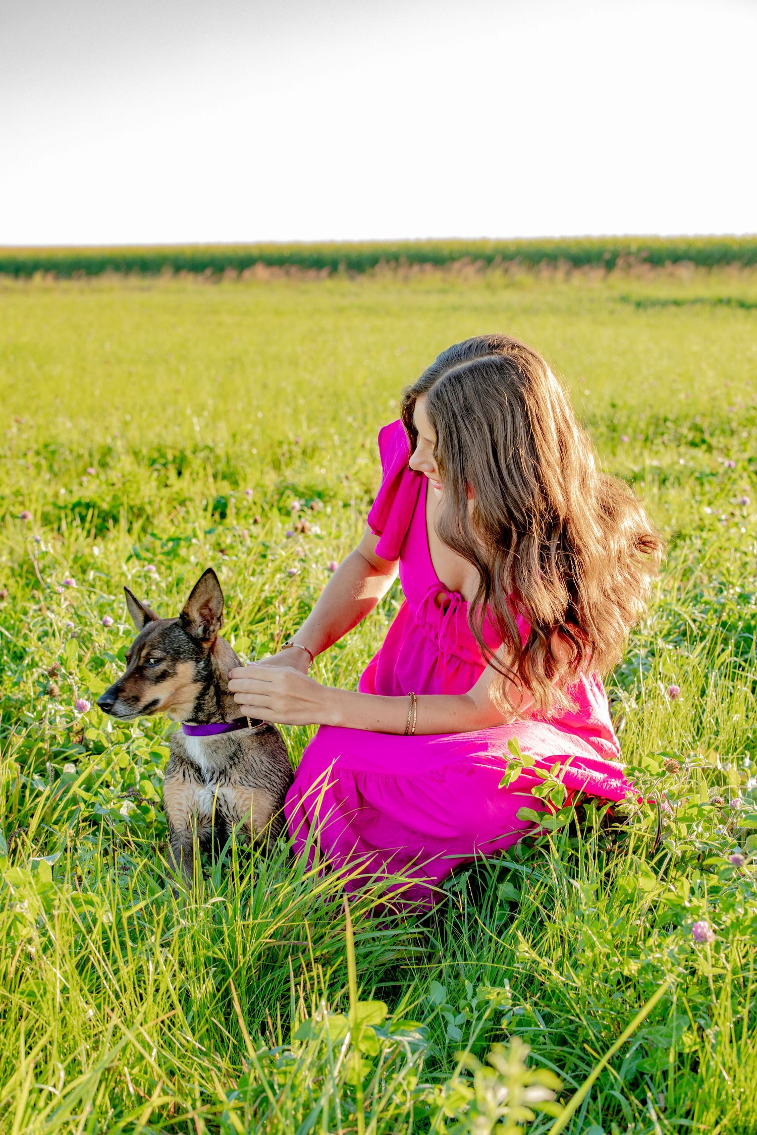 A person in vibrant pink dress lies in a grassy meadow with a dog resting beside them on a sunny day.