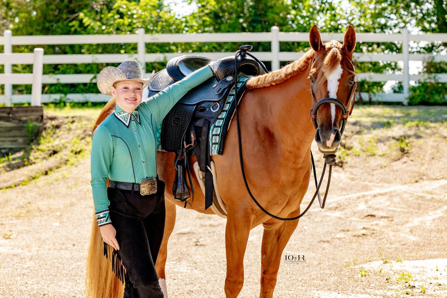 Young equestrian in green shirt and cap stands beside chestnut horse with western saddle near white fence.