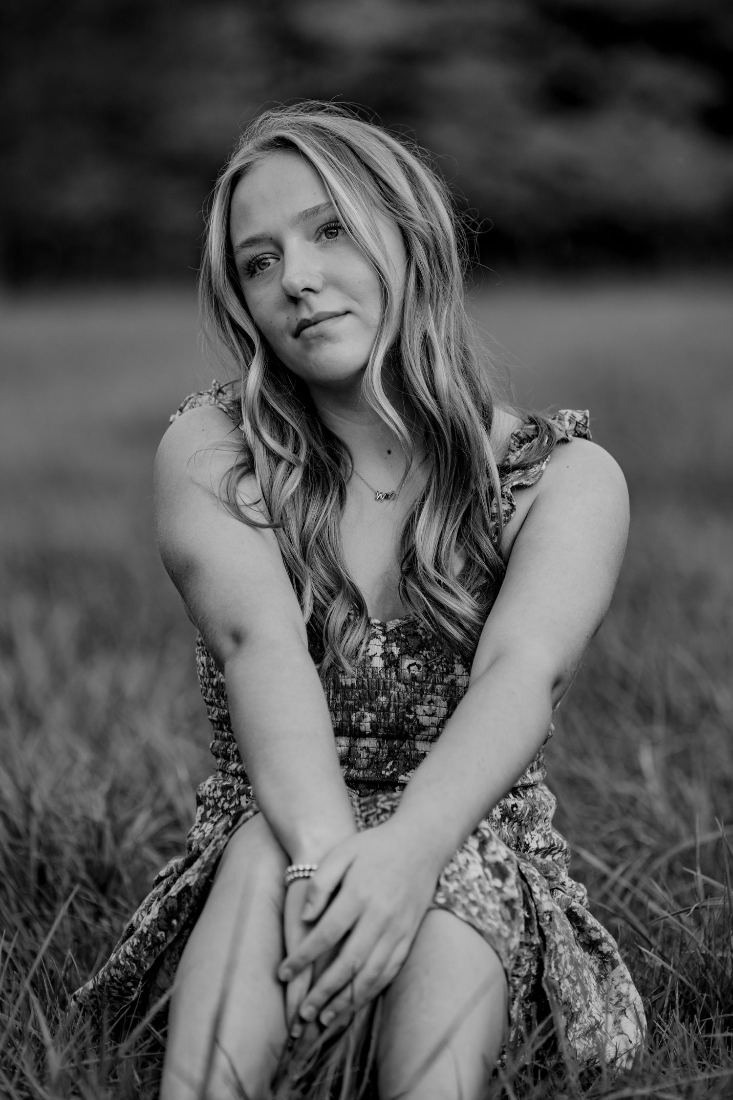 Black and white portrait of someone sitting in a grassy field wearing a floral dress.