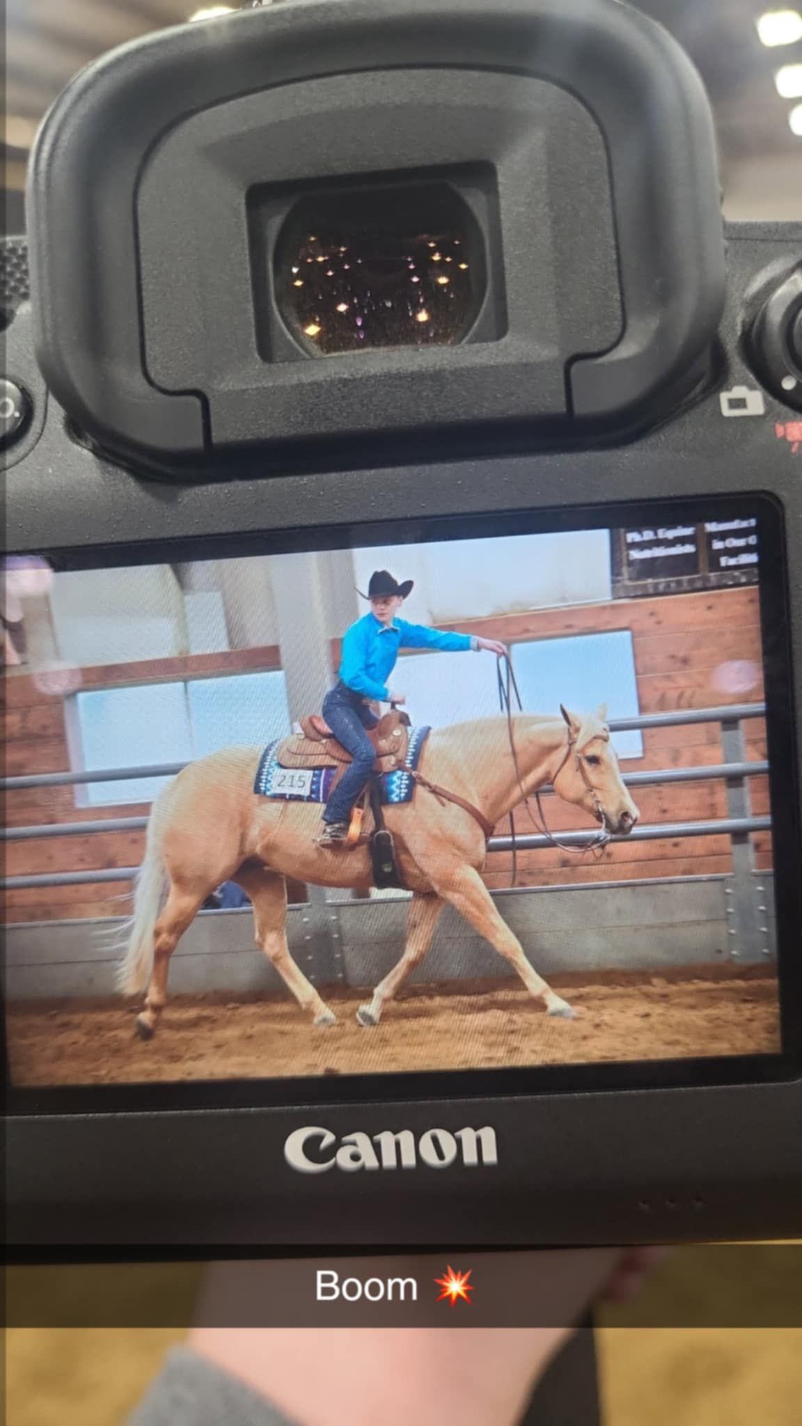 A photographer looking through Canon camera viewfinder at horse rider in blue shirt practicing in arena.