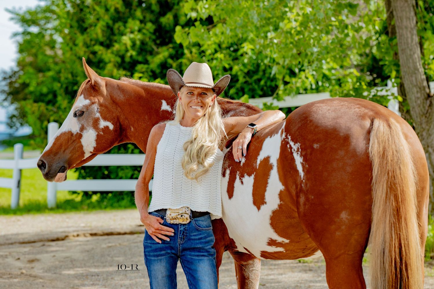 Blonde woman in cowboy hat and jeans poses with a chestnut paint horse by a white fence outdoors.