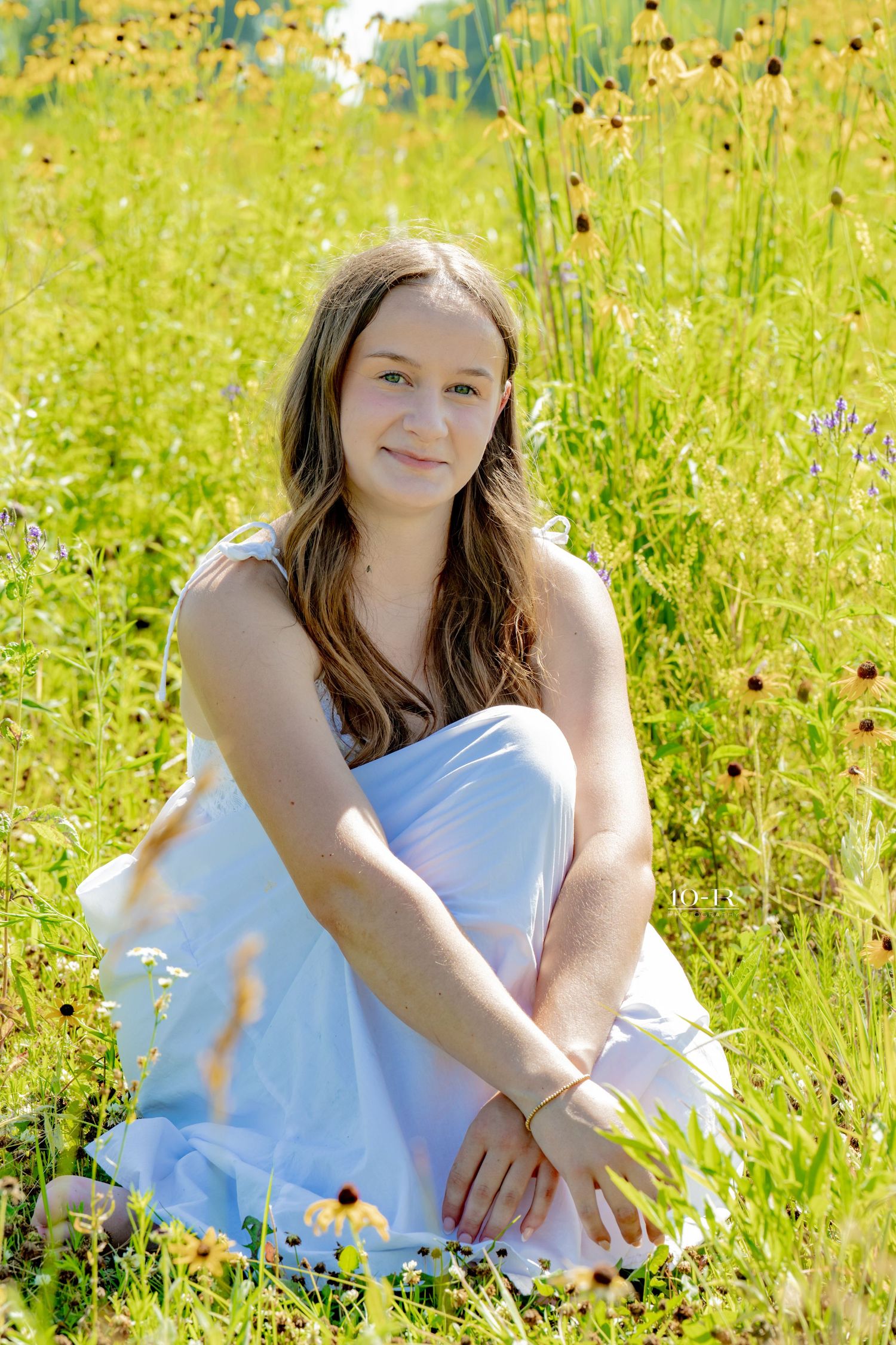 Young woman in light blue dress sitting in a sunny wildflower meadow with yellow flowers.