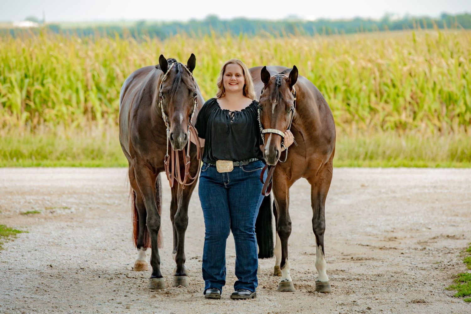 Woman standing between two brown horses on a gravel path with cornfield background.
