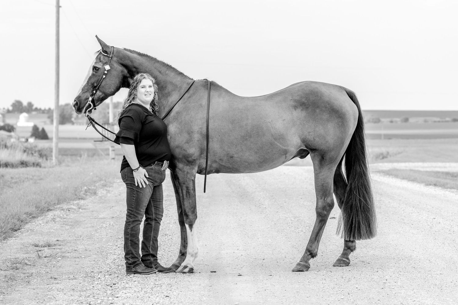 Black and white photo of a woman standing beside a tall horse on a rural gravel road with flat farmland in background.