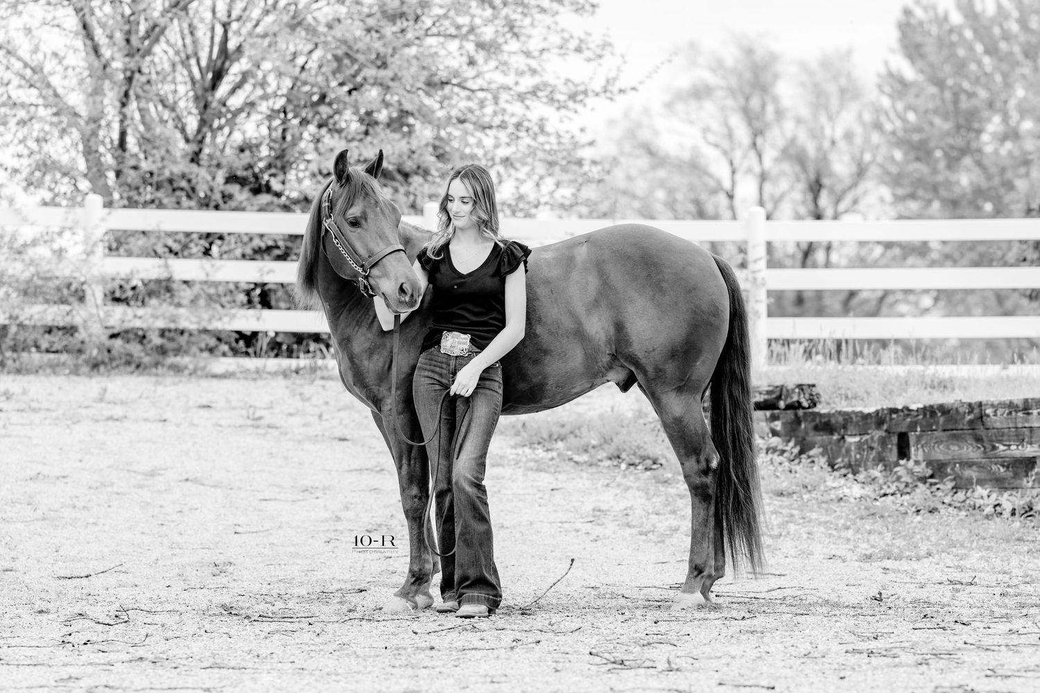 Black and white photo of a young woman standing beside a horse in a fenced ranch setting.