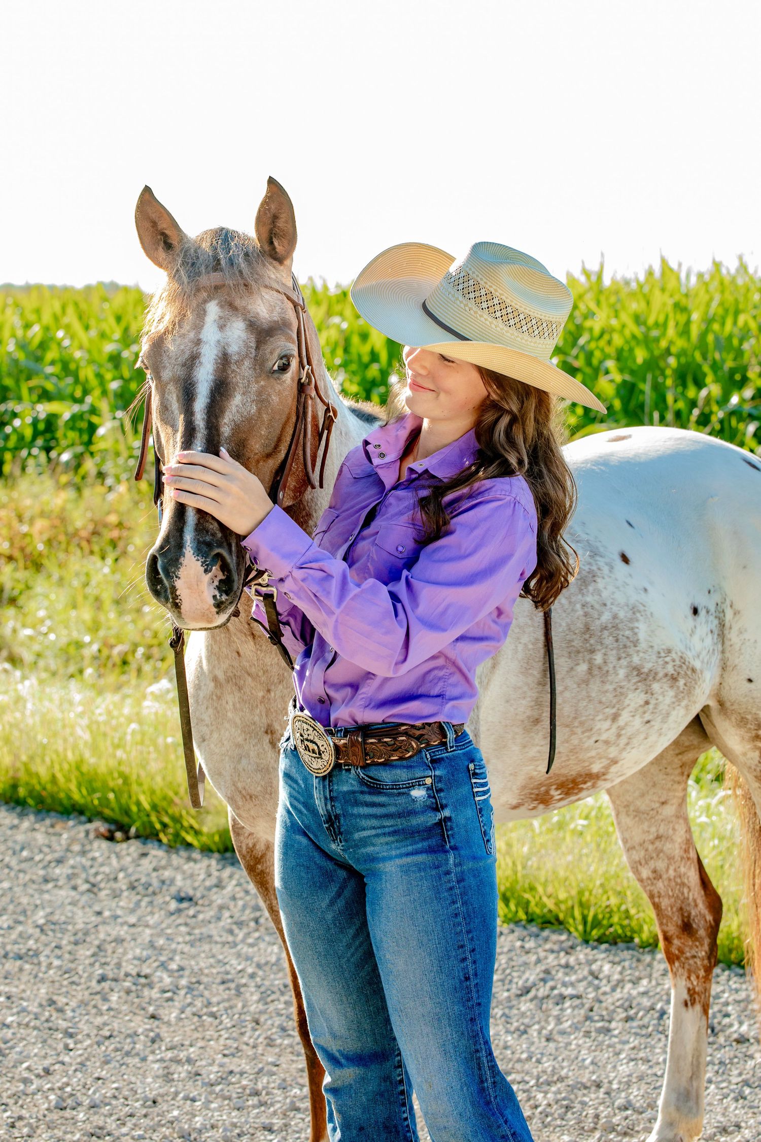 Western style outfit with purple shirt and cowboy hat alongside an appaloosa horse on gravel path.
