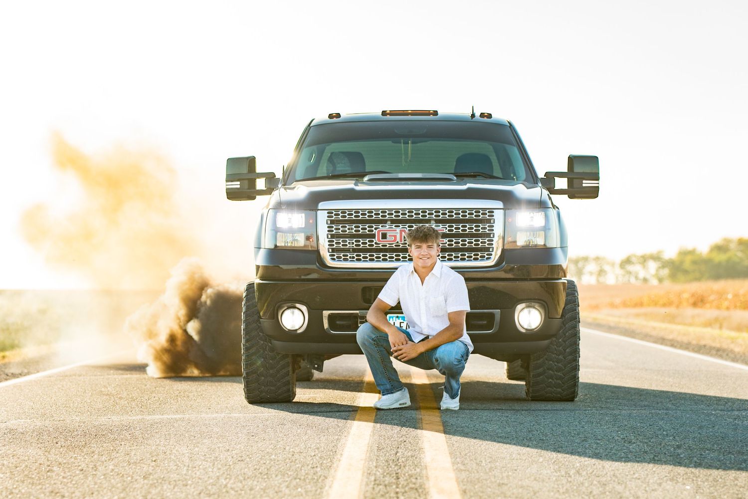 A person sits on the front bumper of a large black GMC truck at sunset on an empty road.