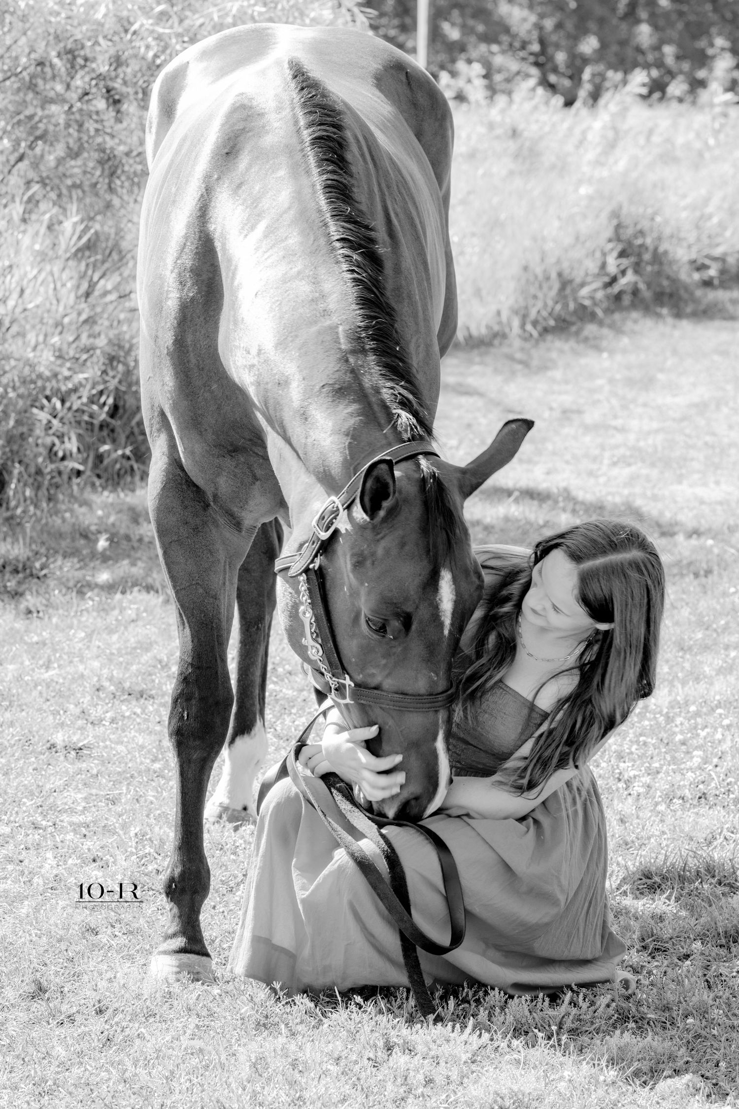 Black and white photo of a woman kneeling on grass, sharing a tender moment with a horse nuzzling her face.