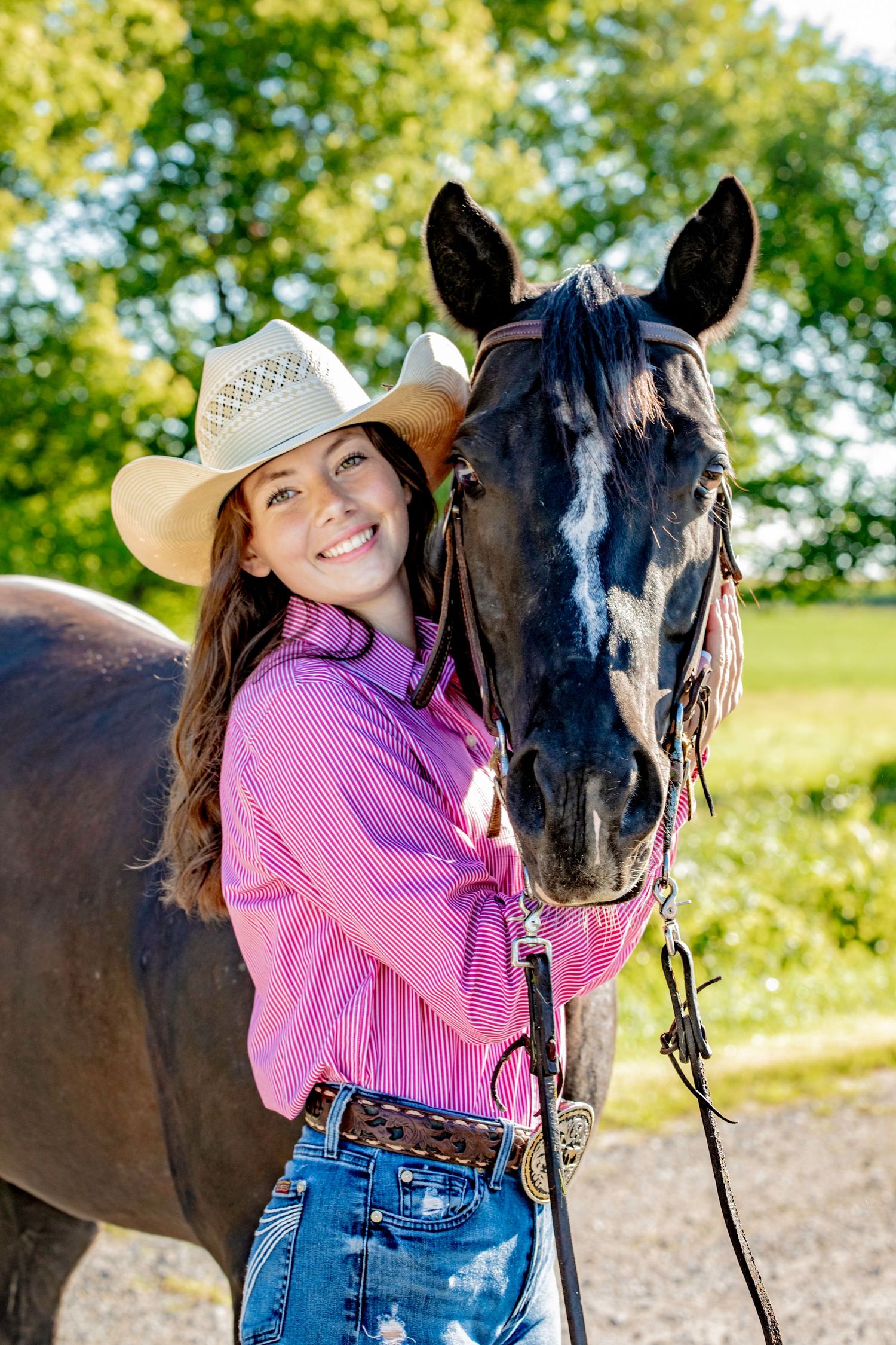 A beautiful brown horse and rider pose together in a sunny field wearing a pink gingham shirt and cowboy hat.