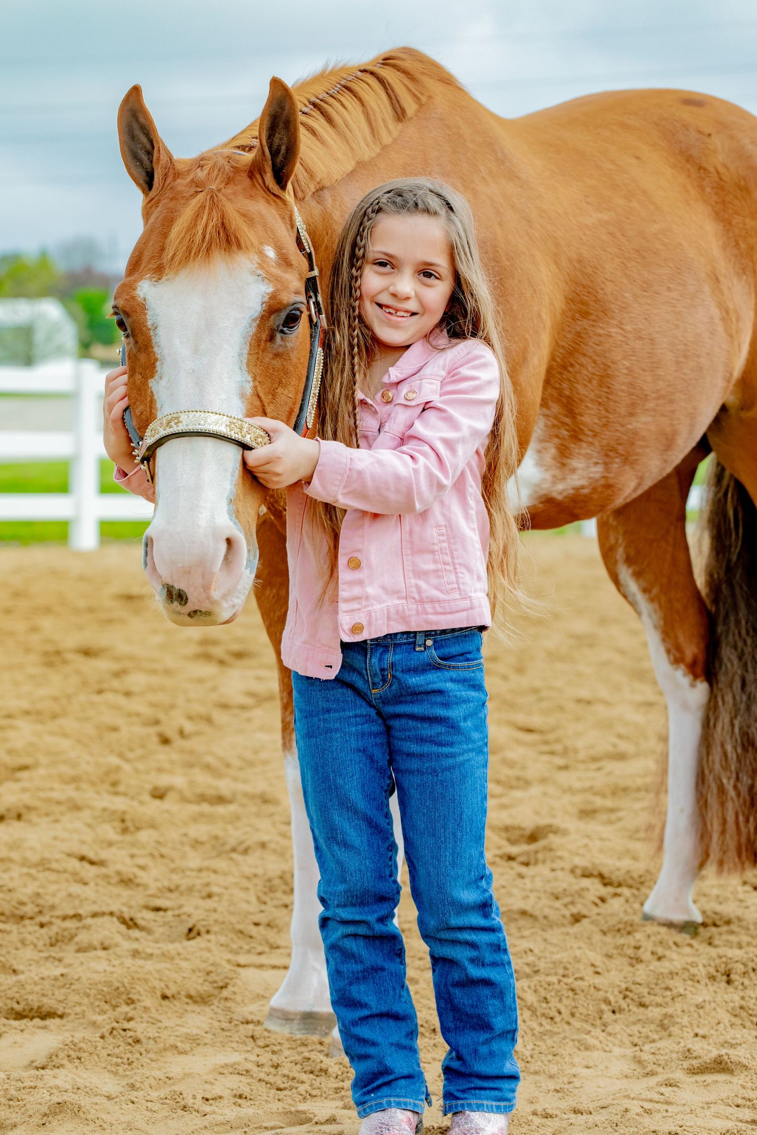 A beautiful chestnut horse with a white blaze stands next to someone in a pink jacket and blue jeans in a sandy riding arena.