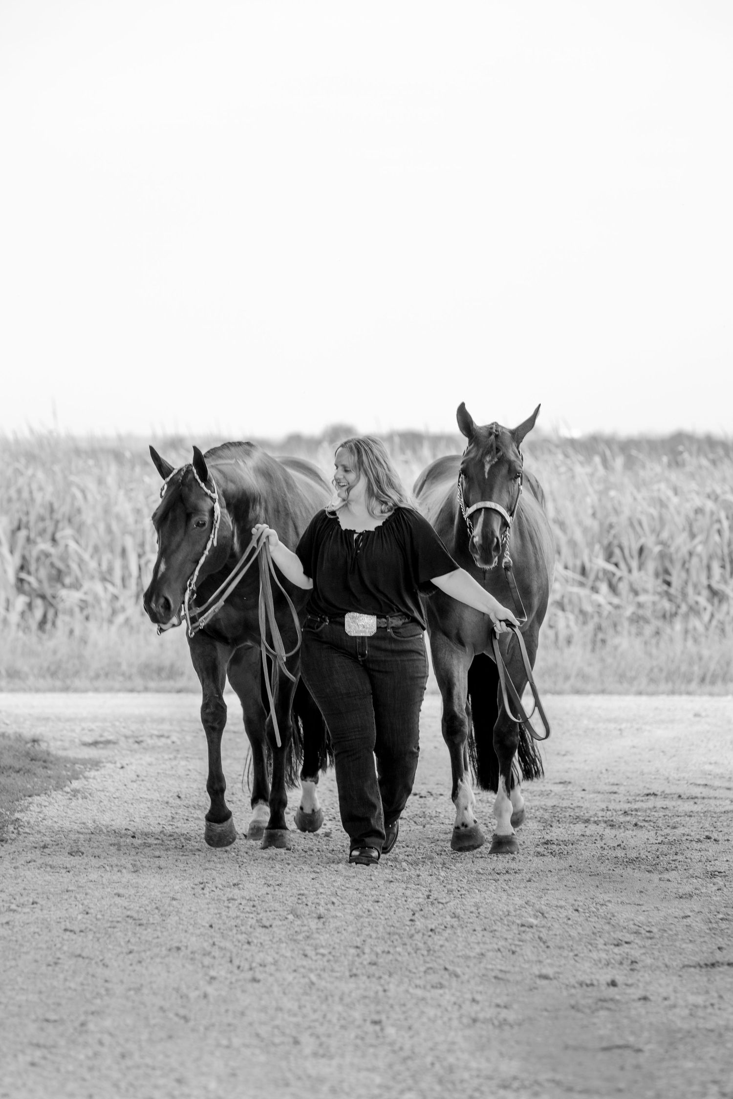 Black and white photo of a woman walking two horses down a dirt path beside a cornfield.