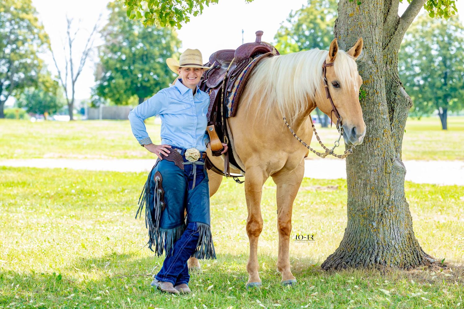 Cowboy standing beside a palomino horse under a tree in a green field, wearing a hat and chaps.