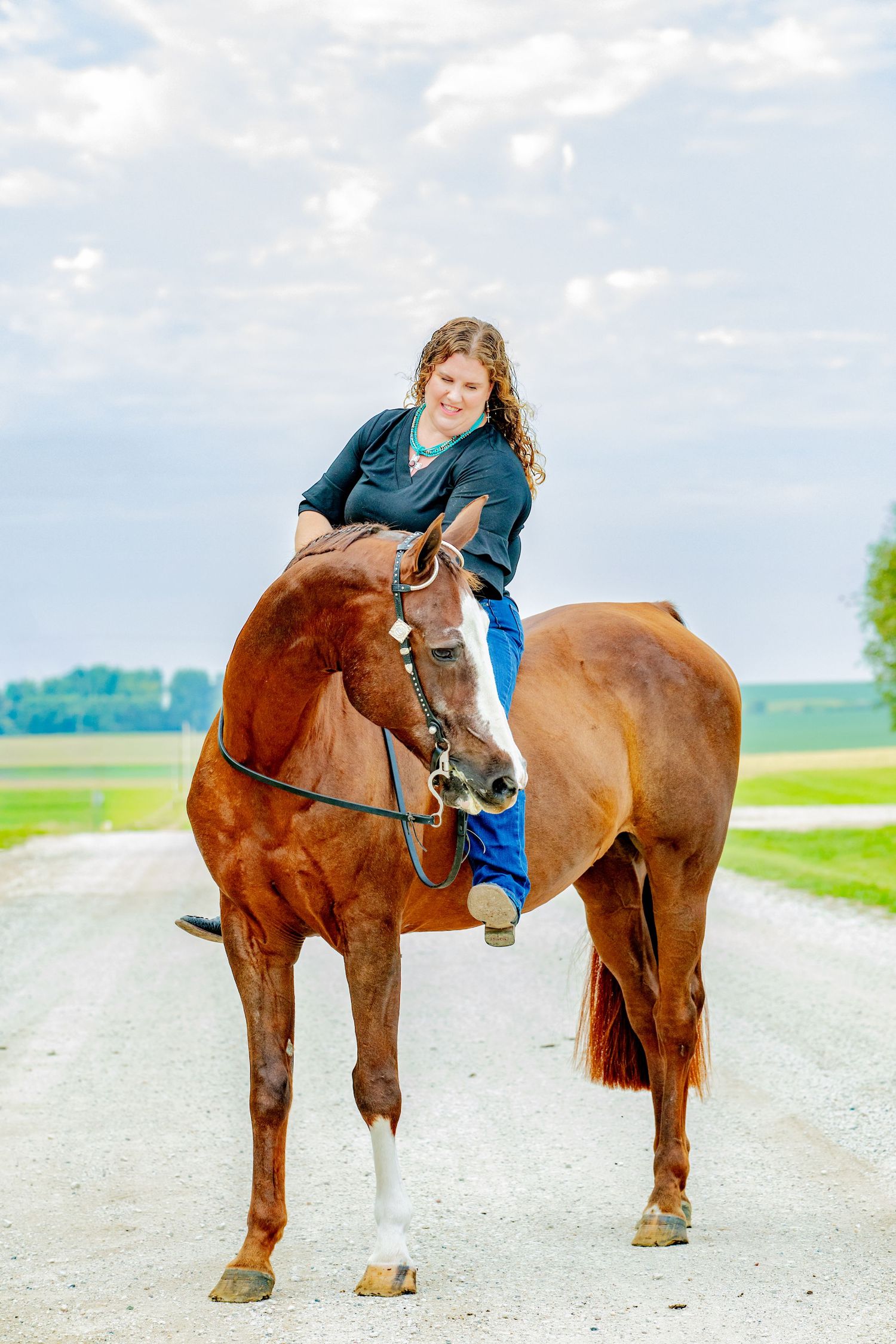 A smiling woman with long curly hair rides a chestnut horse on a gravel road with green fields behind.