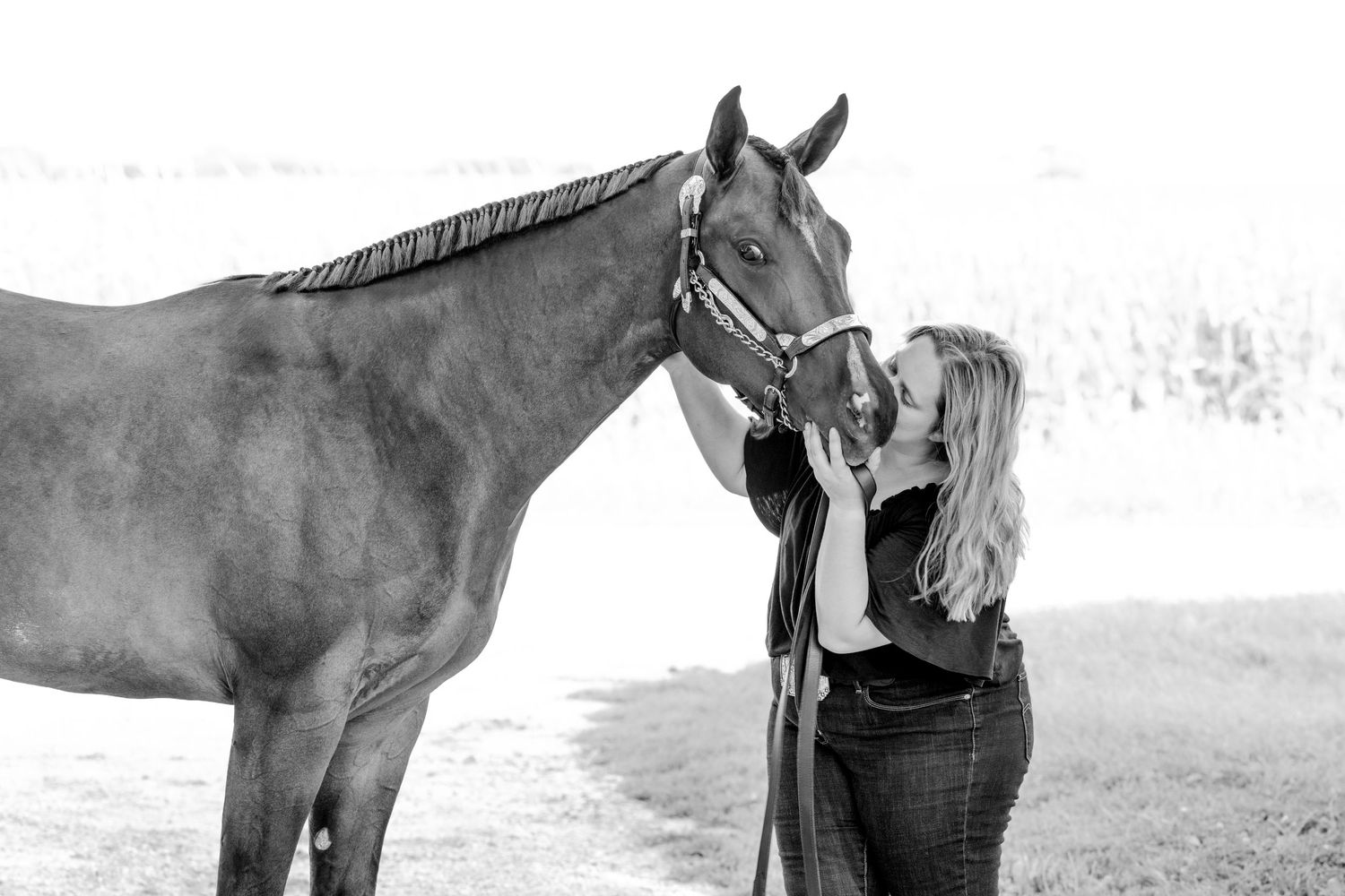 Black and white photo of a woman kissing a brown horse wearing a halter in an outdoor field setting.
