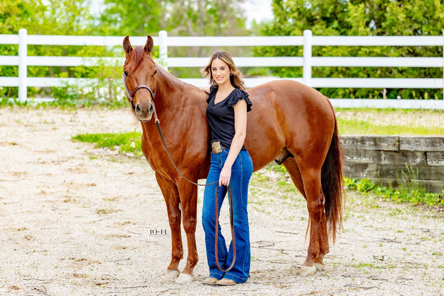Young woman in jeans and black top poses with a chestnut horse by a white fence in an outdoor arena.