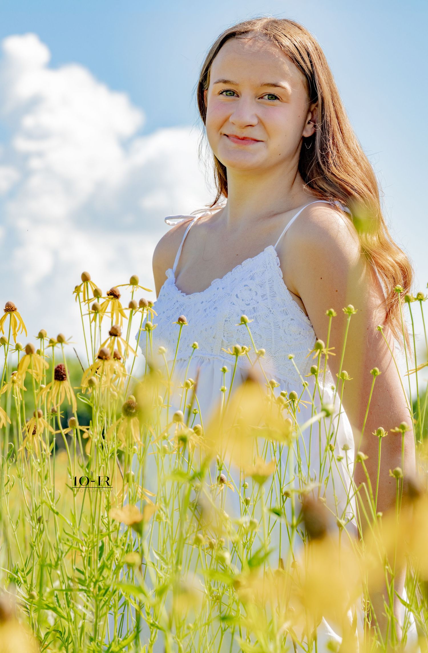 Young woman in white sundress smiling among wildflowers in a sunny field with blue sky.