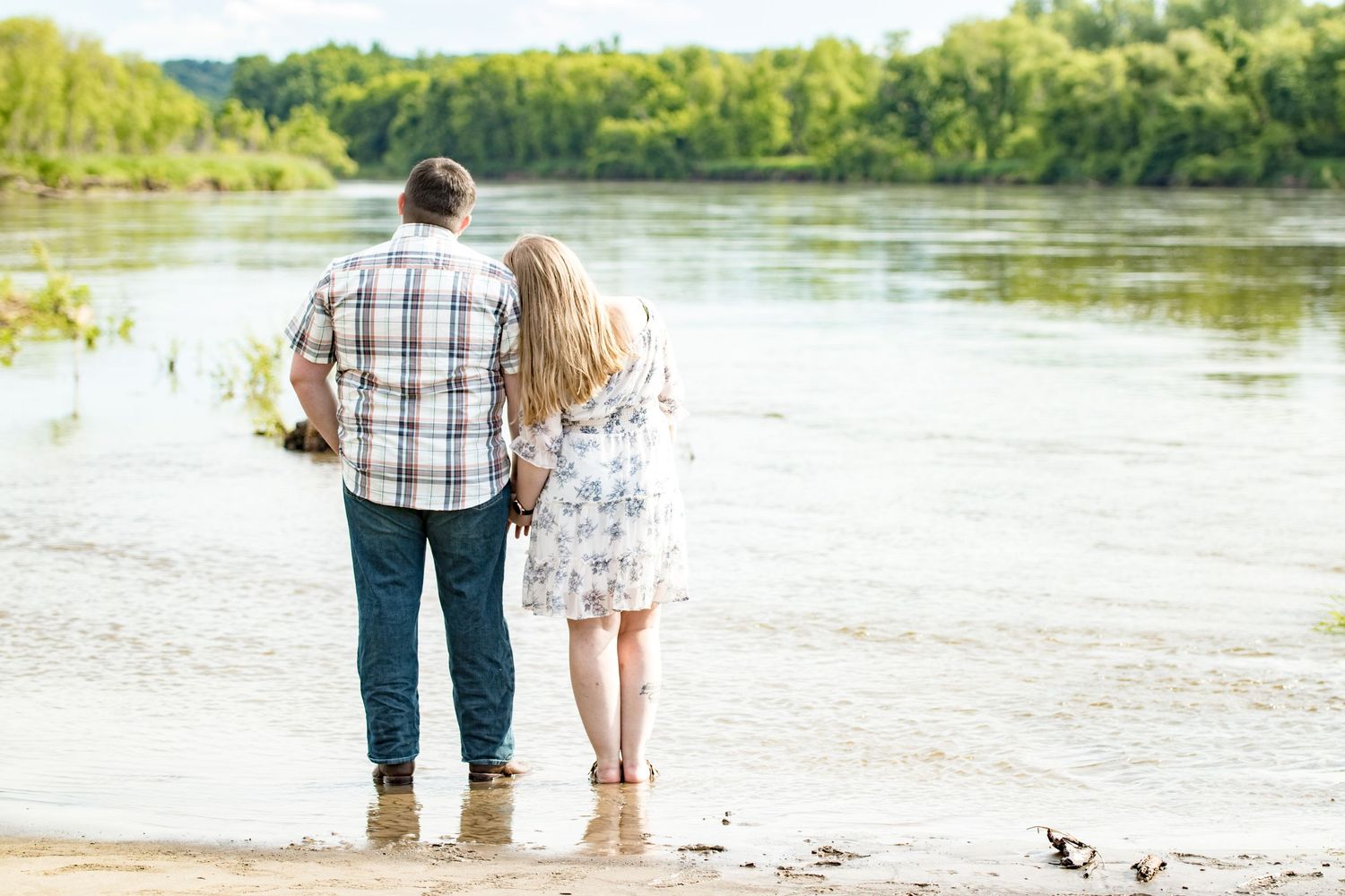 A couple standing together at the edge of a serene lake surrounded by trees.