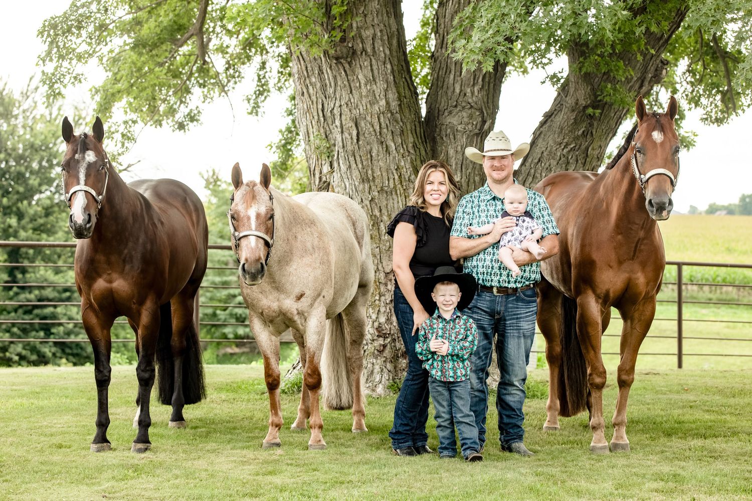 Family portrait with four horses standing under a large tree in a rural setting.