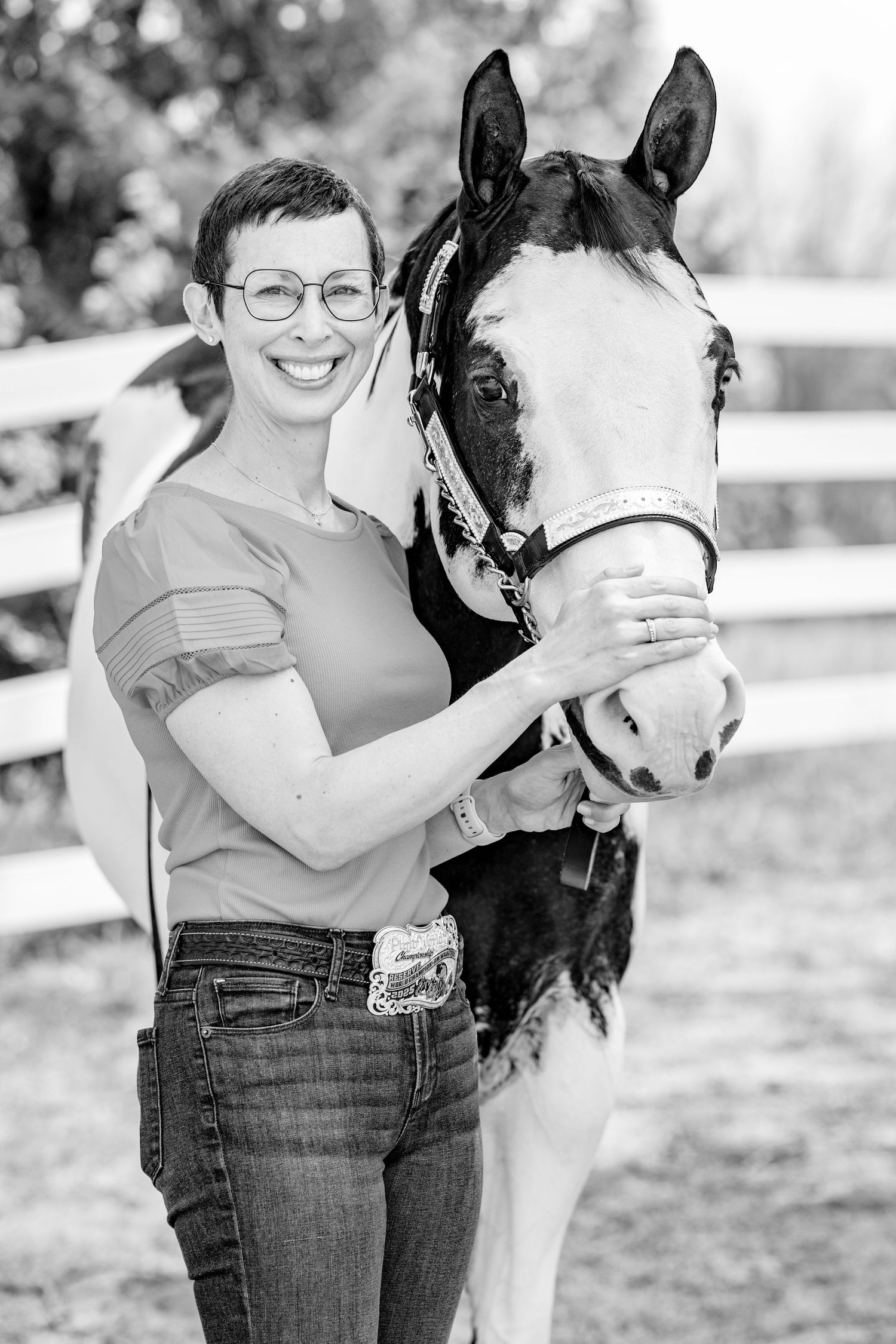 Black and white photo of a smiling woman with short hair and glasses posing with a paint horse near a white fence.