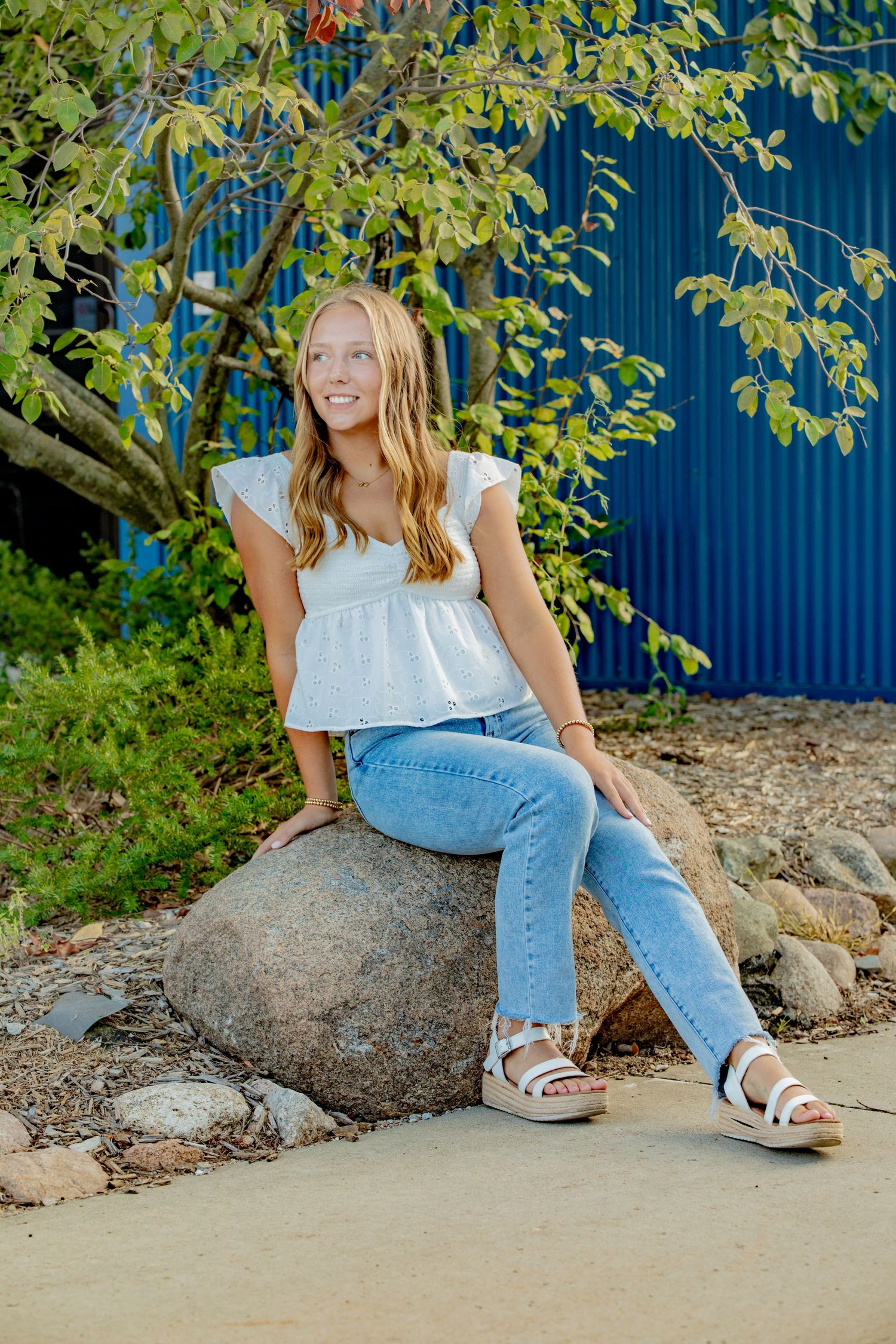 Woman in casual white top and denim relaxes on large rocks against blue wall with green foliage.